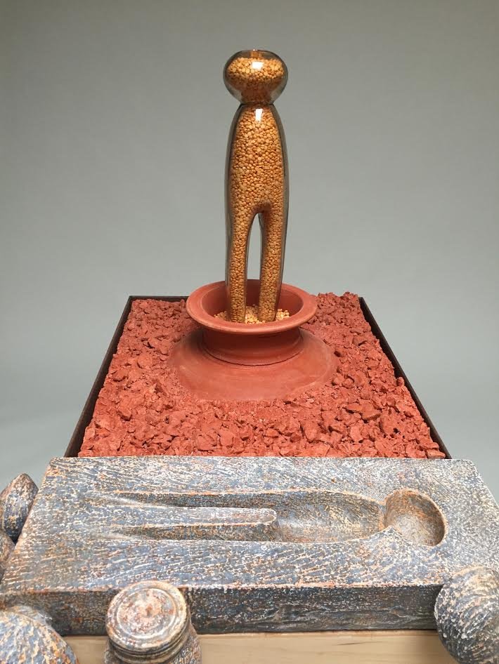 A sculpture of a Mother Pin made from a glass container filled with tiny yellow objects, standing on a circular base within a reddish dish, on a textured red surface, with a rusted metal object in the foreground.