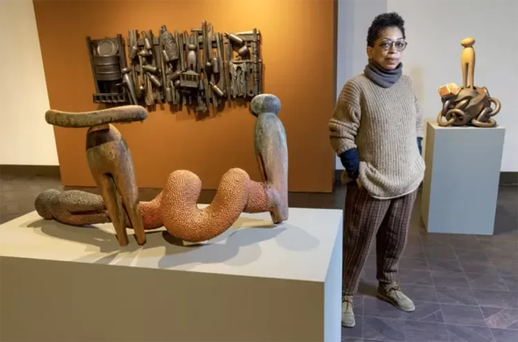 Artist, Syd Carpenter, standing in an art gallery next to abstract sculptures, with one sculpture resembling a snake and another resembling a chair, and wall-mounted art pieces in the background.