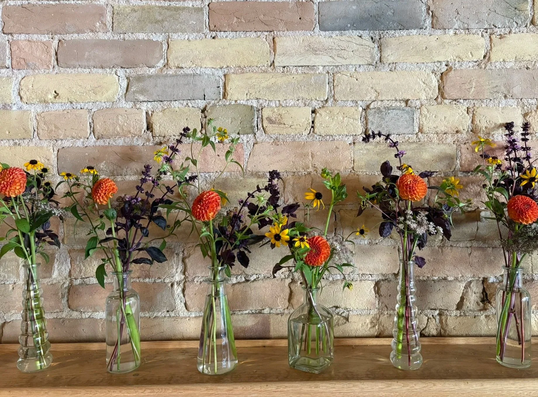 Five vases with colorful flowers on a wooden surface against a brick wall background.