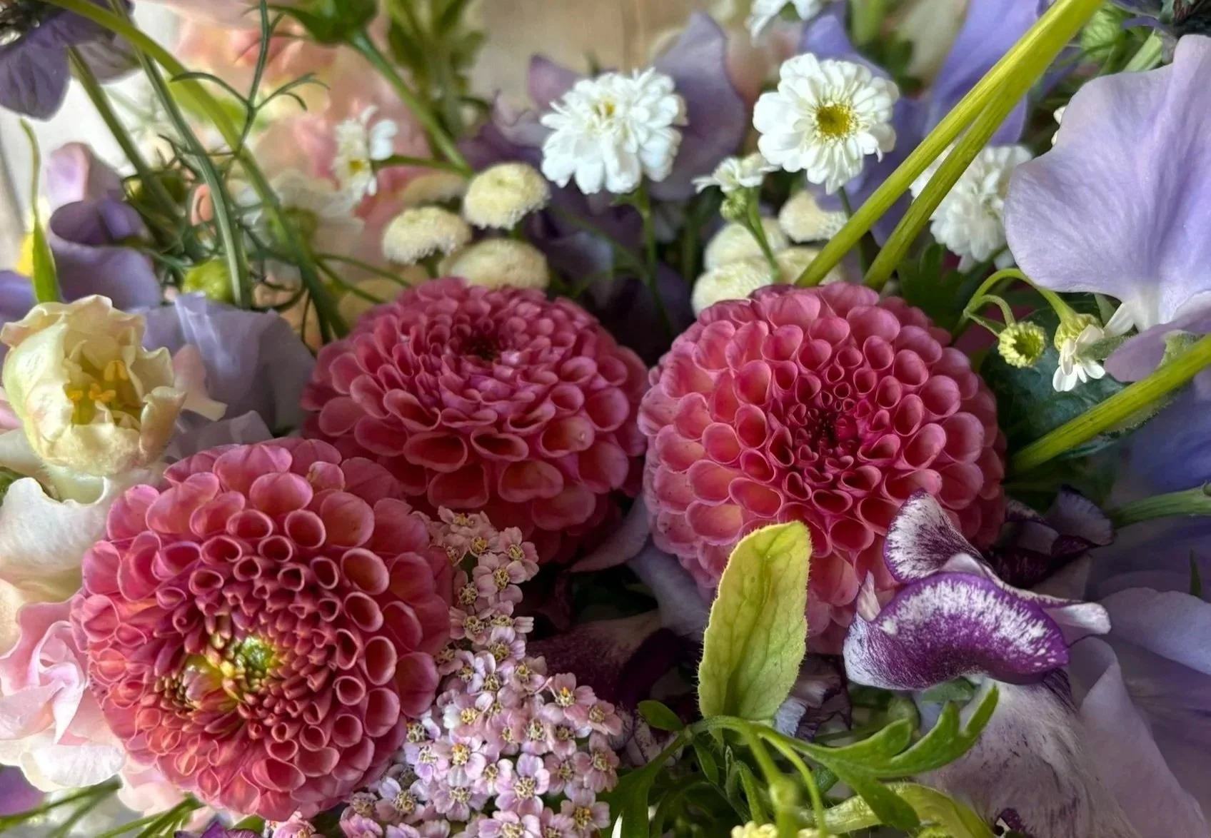 Close-up of a colorful flower arrangement with mauve dahlias, purple sweetpeas, white feverfew, and various green leaves.
