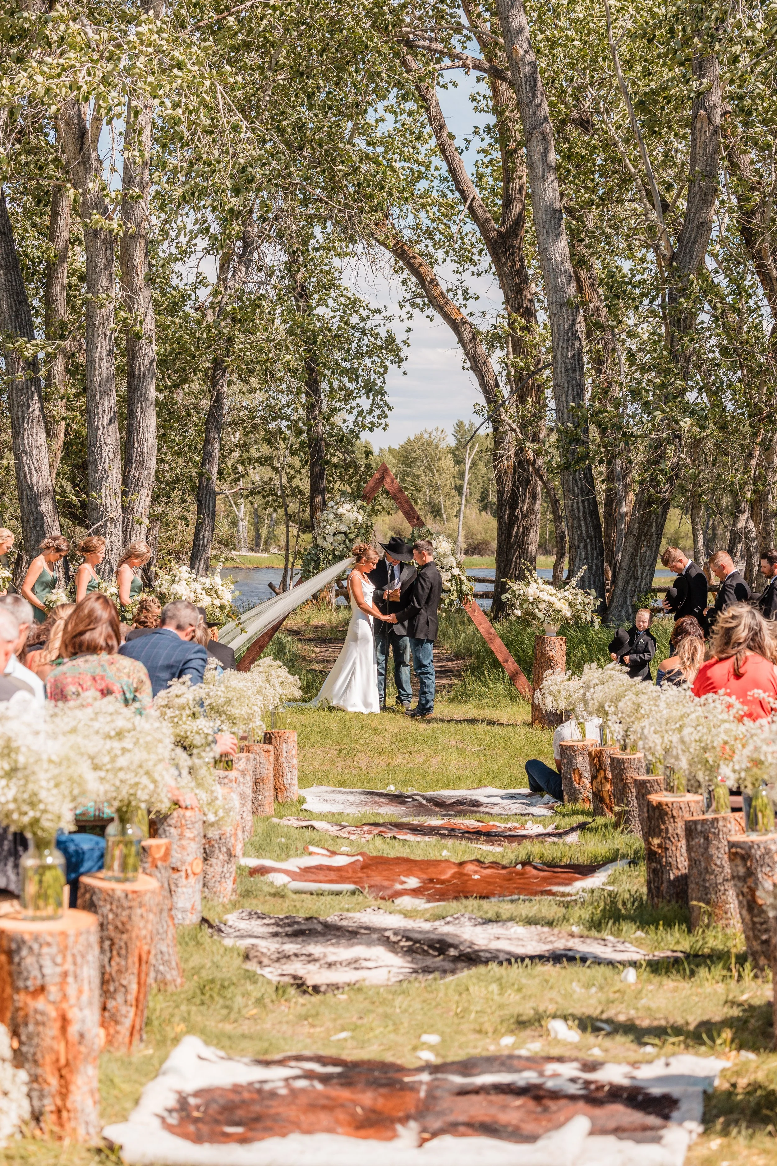 Outdoor wedding ceremony in a forested area near a river with guests seated, vases full of baby's breath on logs pedestals at the end of each row of seats, a couple exchanging vows, and an officiant standing with them.