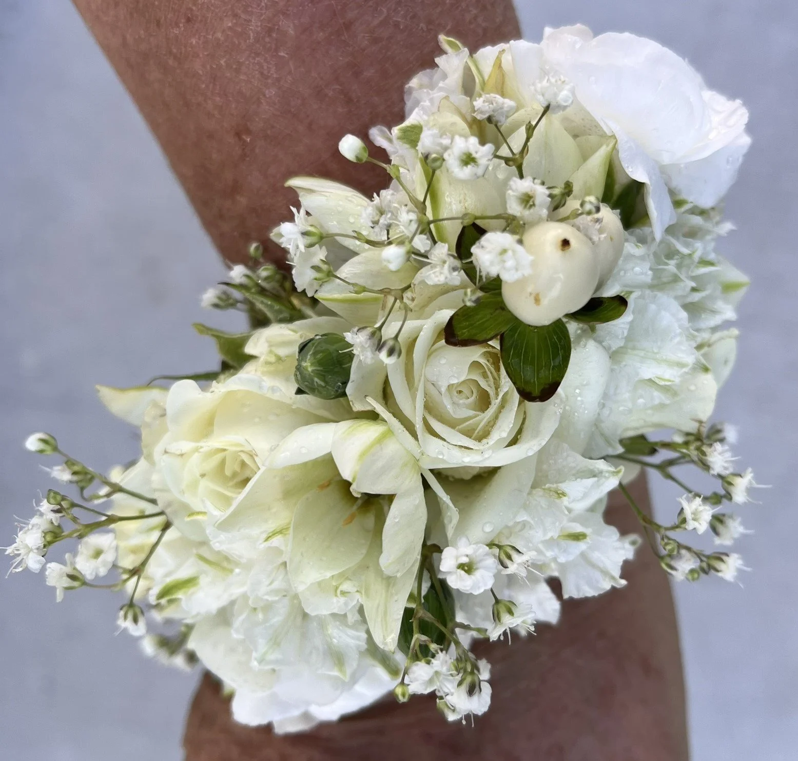 A person's arm holding a white floral boutonniere with roses, baby's breath, and other white flowers, with water droplets on the petals.