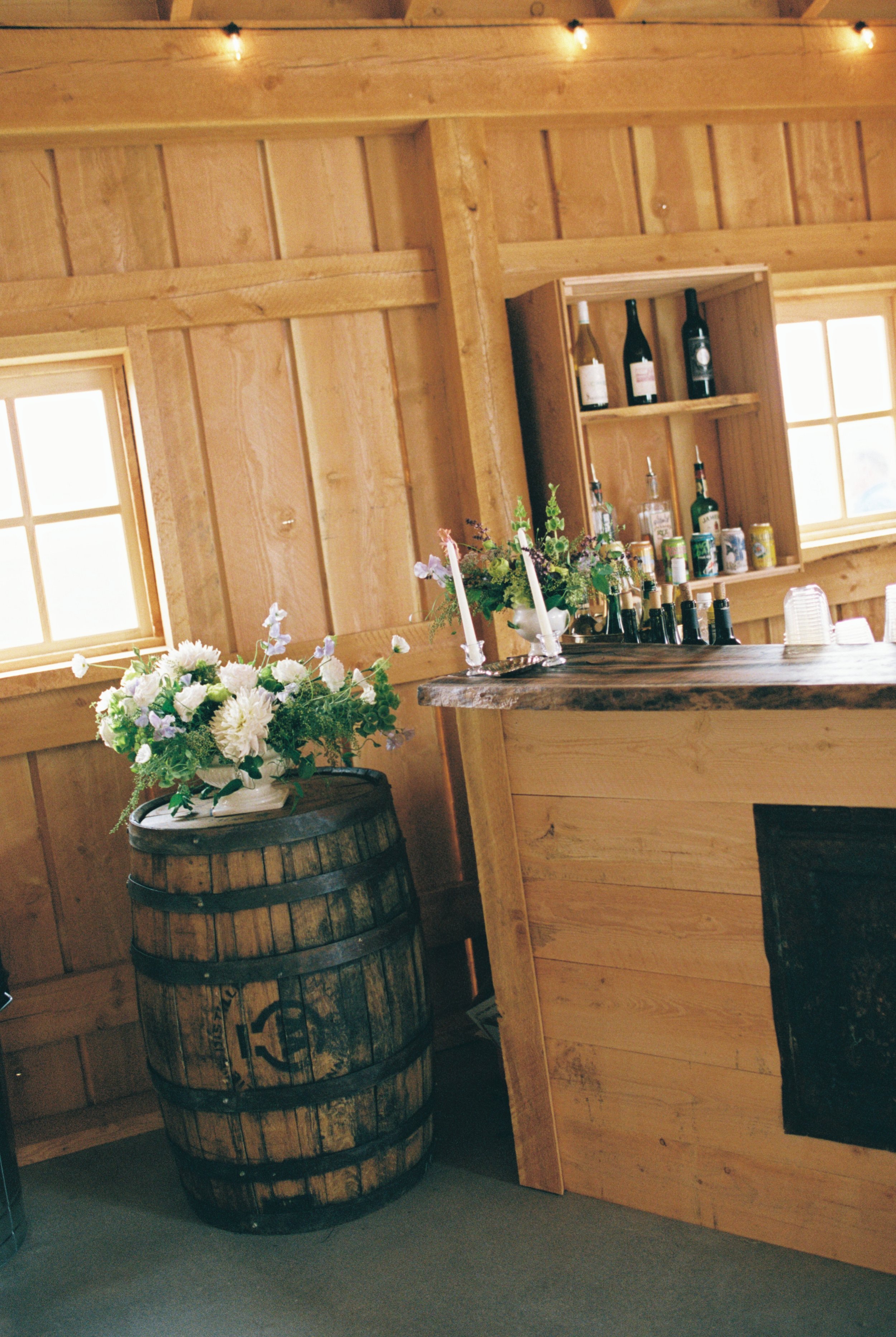 Interior of a wooden bar with a flower arrangement on a barrel, bottles of alcohol on shelves, and soft lighting.