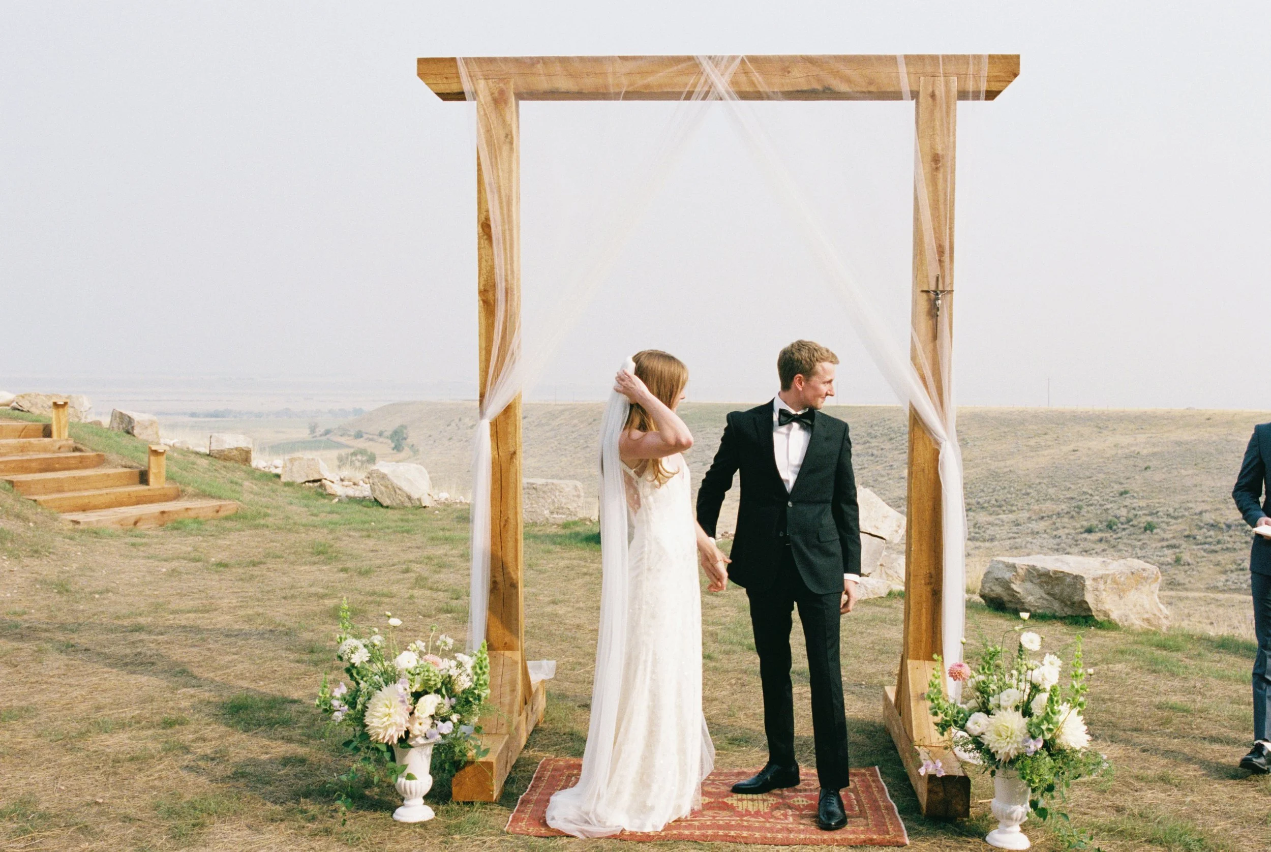 A bride and groom holding hands under a wooden wedding arch outdoors, with large flower arrangements at the base, in a scenic, open landscape.