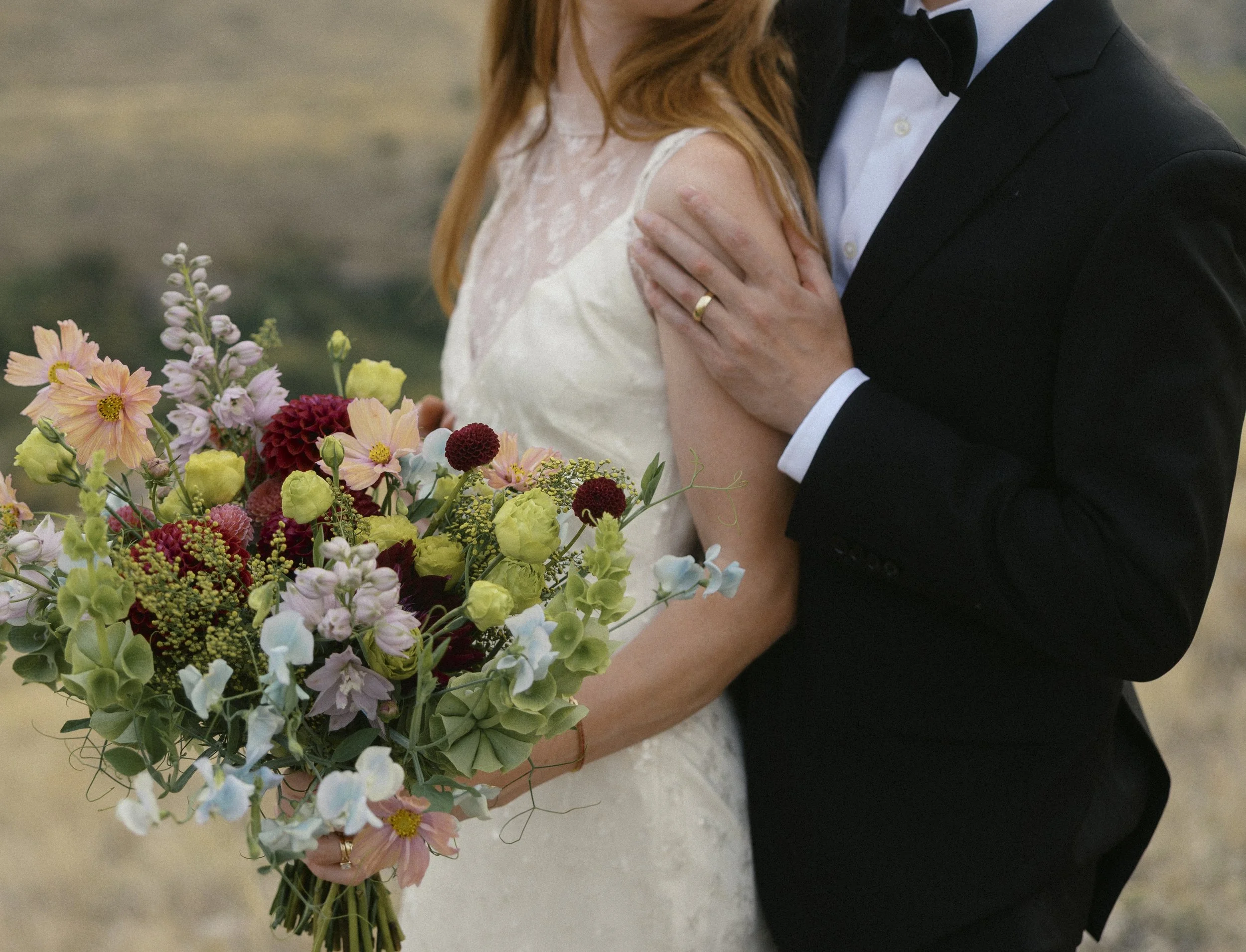 Close-up of a bride and groom on their wedding day. The bride is holding a colorful bouquet of flowers, and the groom is wearing a black tuxedo with a bow tie, holding her close.