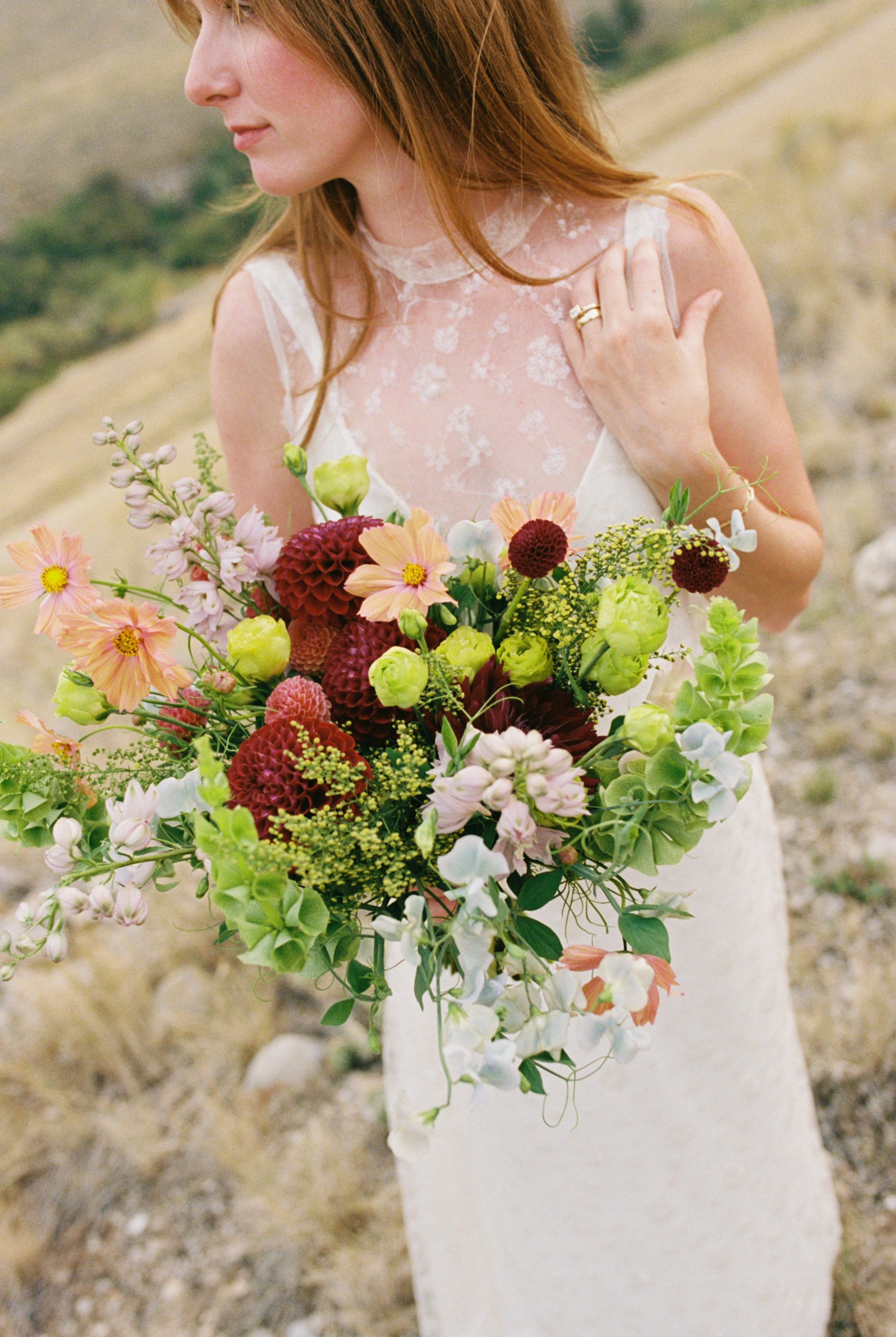 A bride in a lace wedding dress holding a bridal bouquet of flowers outdoors.
