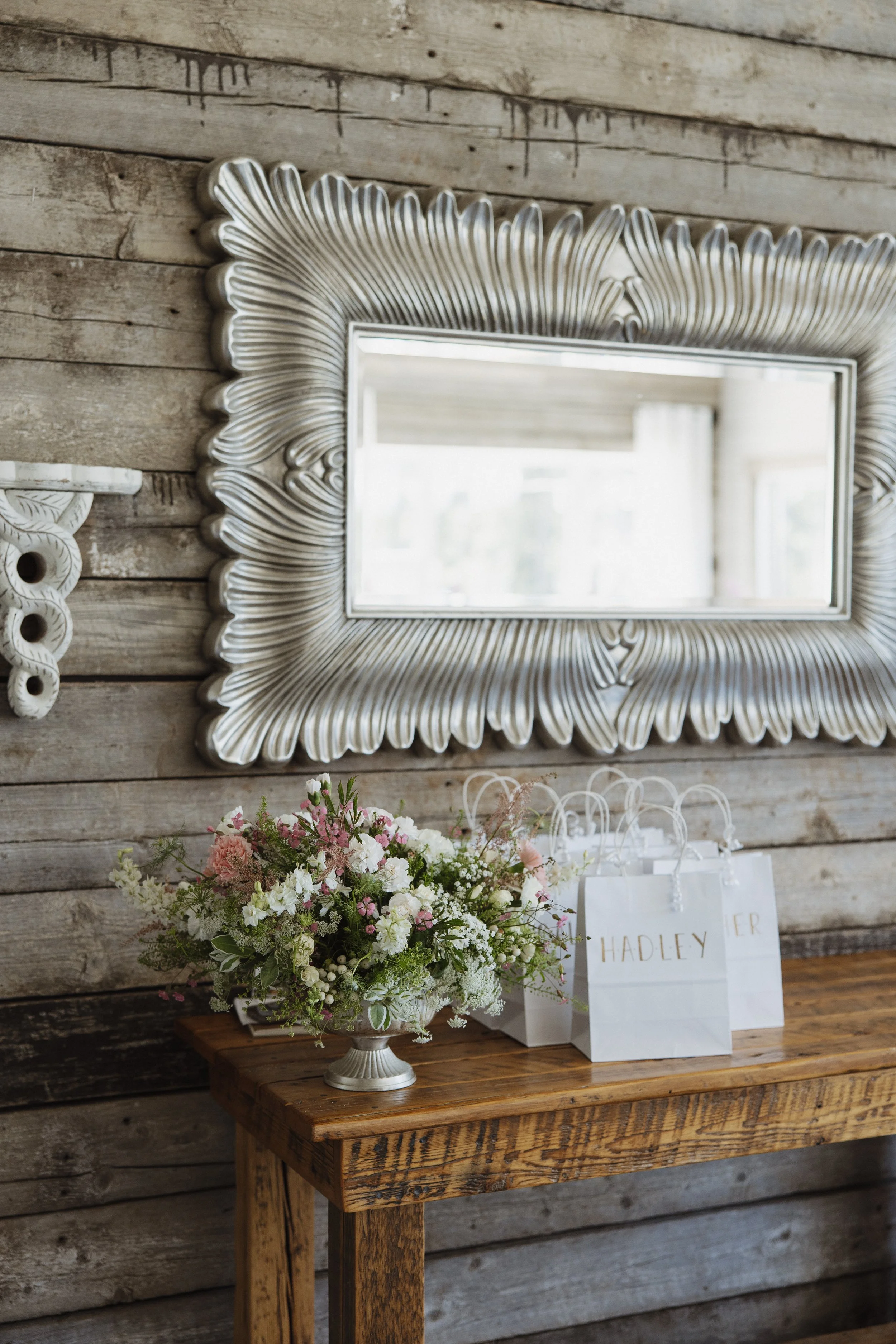 A decorative silver-framed mirror hanging on a weathered wooden wall above a wooden table. The table holds a white and green floral arrangement and white gift bags labeled with names.