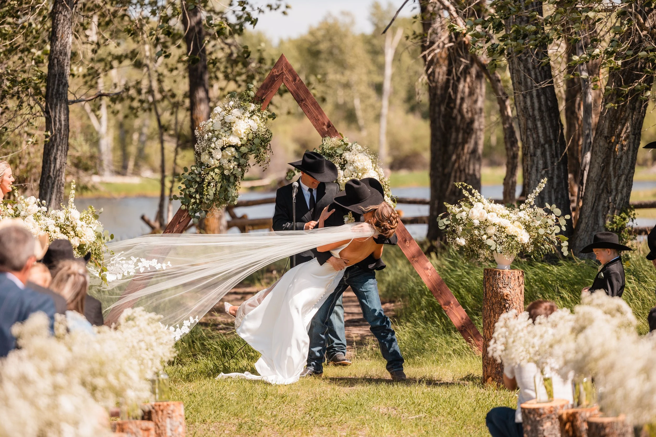 A wedding ceremony taking place outdoors by a lake with trees, decorated with flowers and a wooden triangle arch, where a bride in a white dress is with a groom in a black cowboy hat.