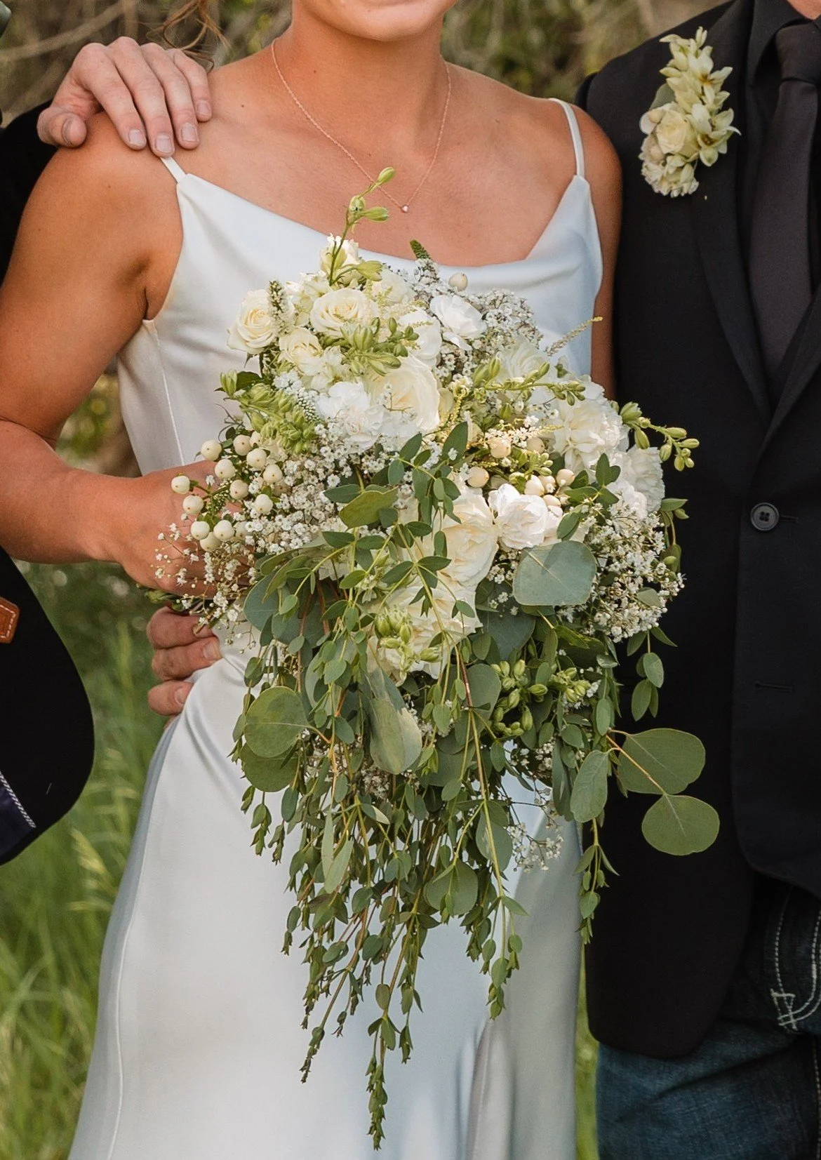 Close-up of a bride holding a cascading bouquet of white roses, other white flowers, and eucalyptus, with a groom partly visible, both dressed in wedding attire outdoors.