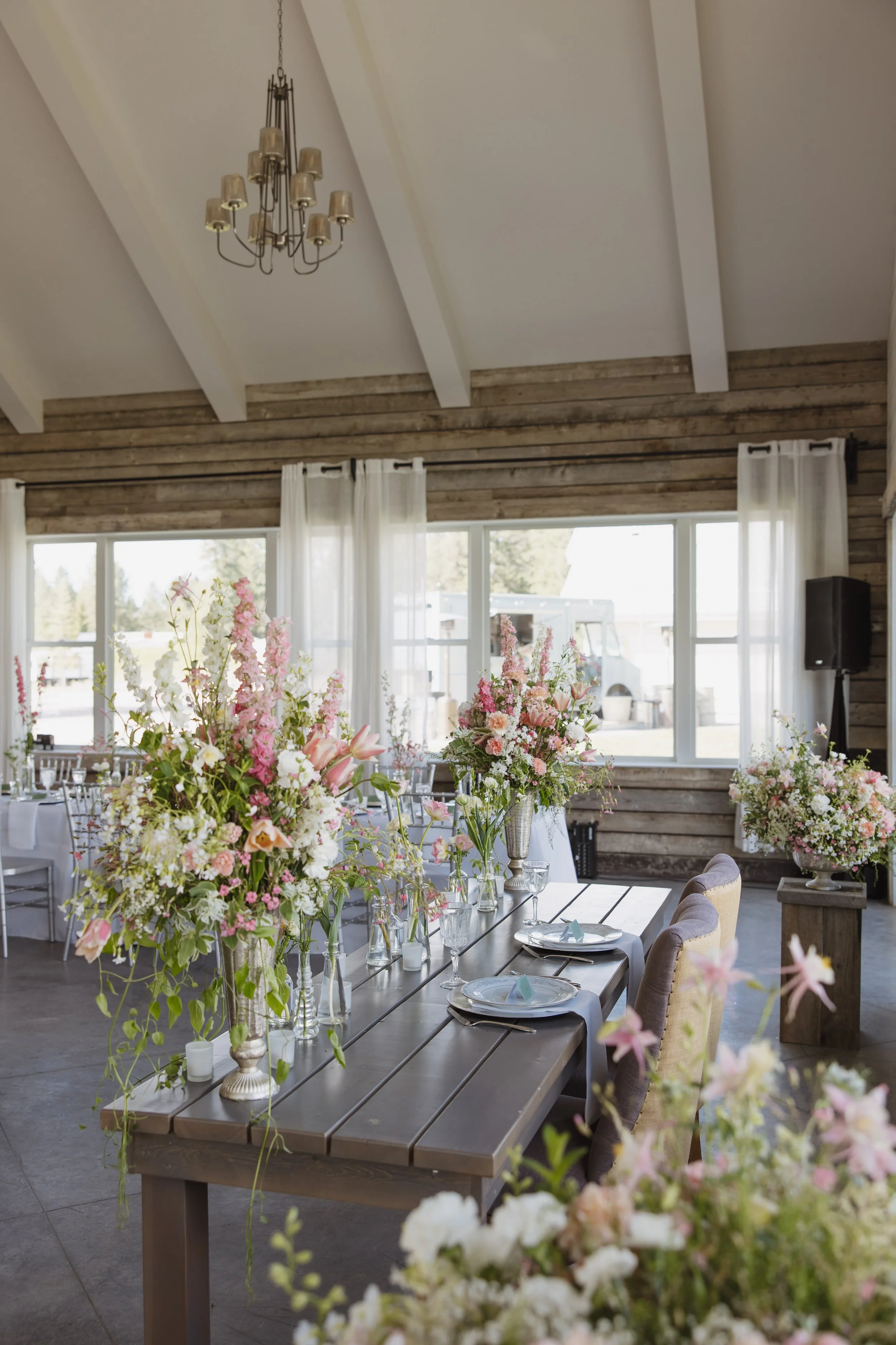 A decorated dining area with floral centerpieces on a rustic wooden table in a bright room with large windows, white curtains, and a chandelier.