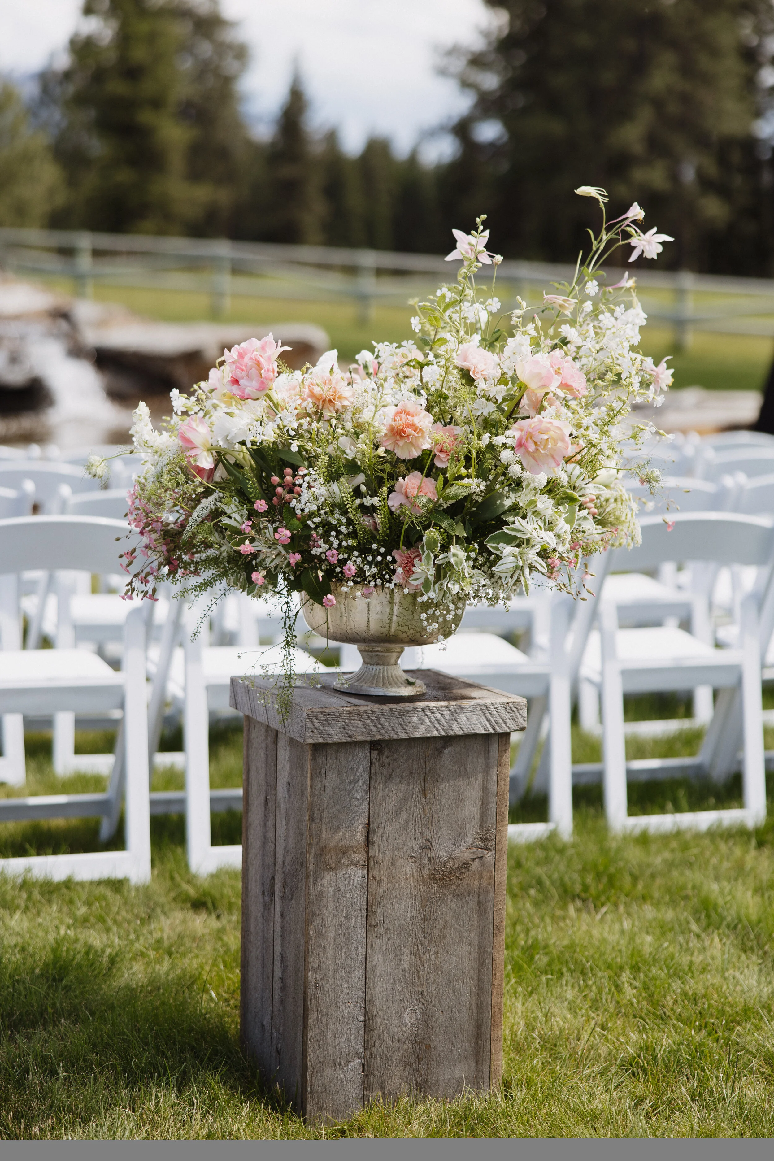 A large floral centerpiece with pink, white, and light purple flowers in a white urn, placed on a rustic wooden stand at an outdoor event setting with white chairs in the background.