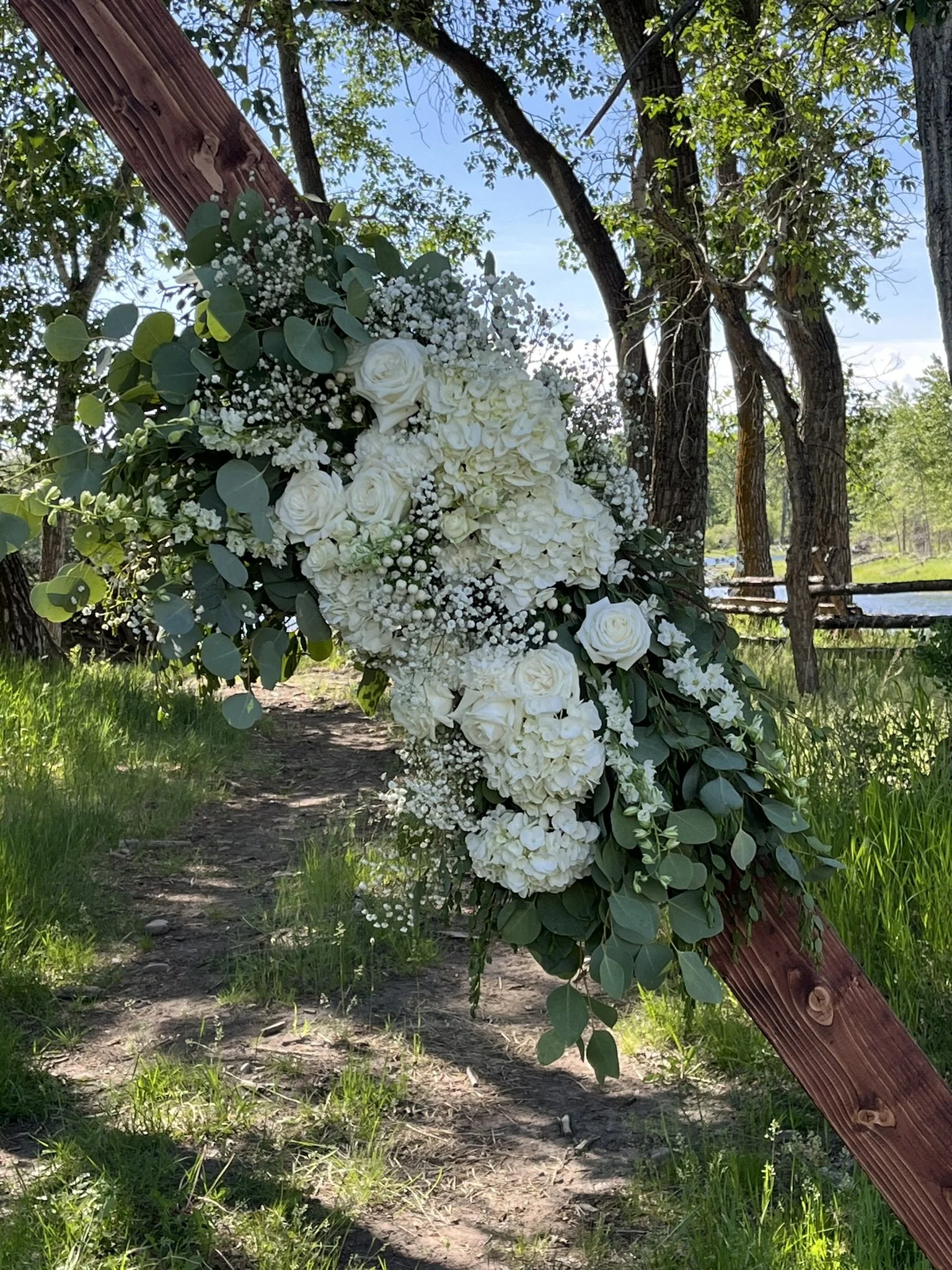 A floral arrangement with white roses, hydrangeas, baby's breath, and eucalyptus leaves attached to a wooden support on a nature trail.