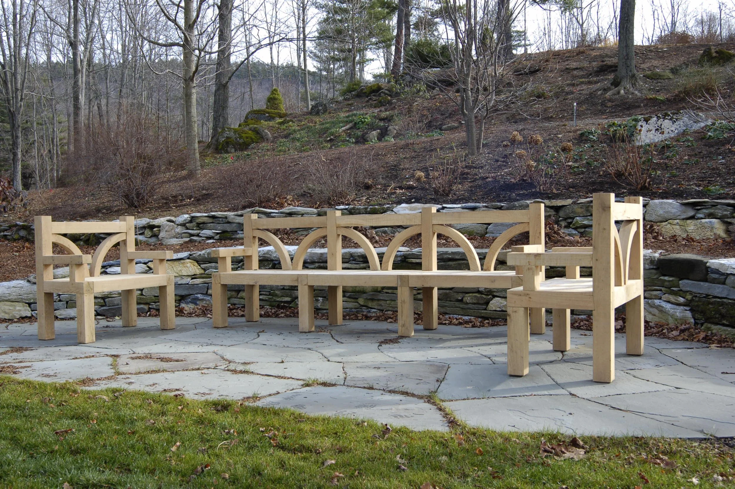 Wooden outdoor bench on paved stone patio with a natural wooded background and a stone retaining wall.