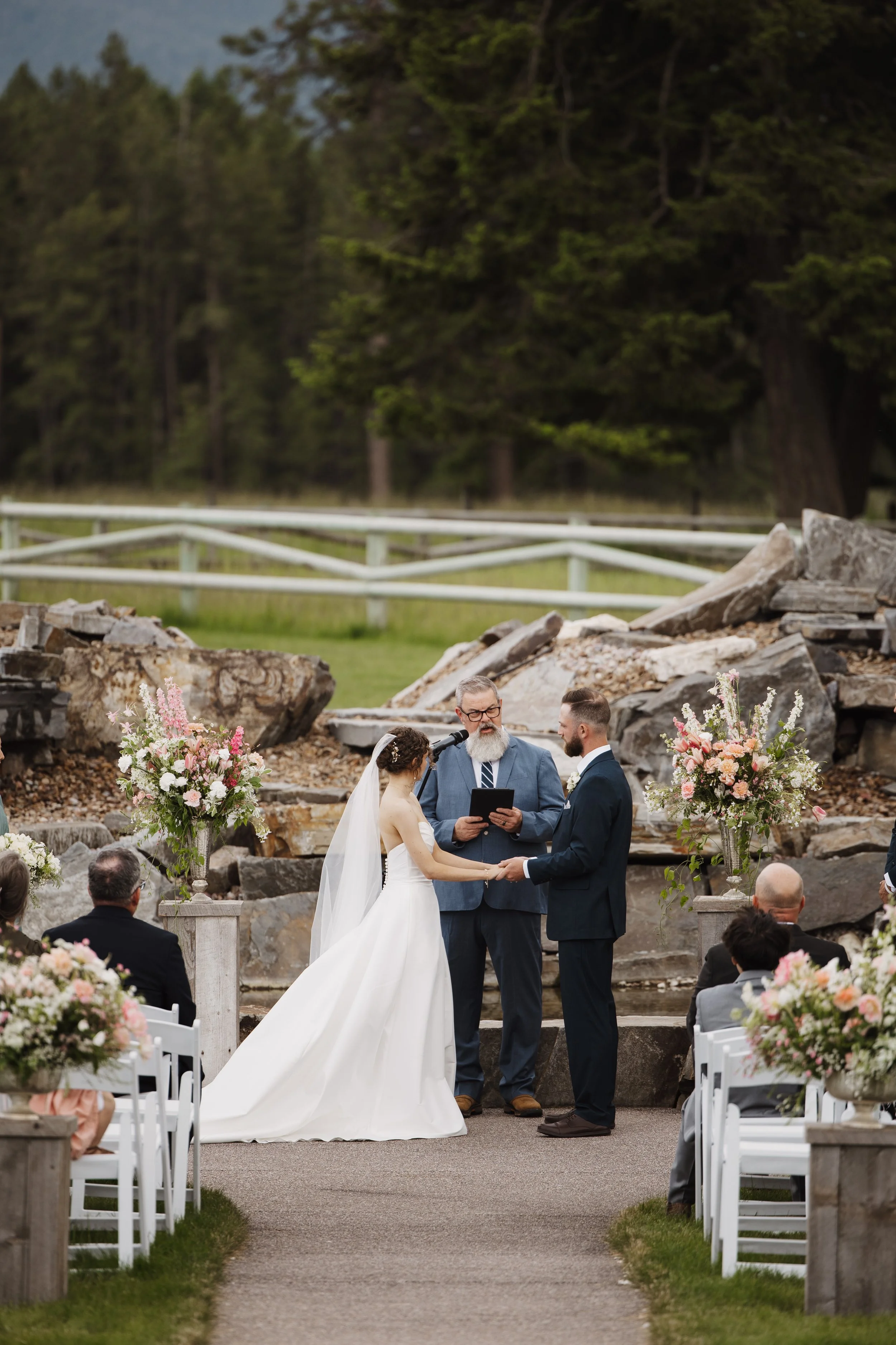 A couple getting married outdoors during a ceremony, with an officiant reading vows, surrounded by floral arrangements, seated guests, and nature in the background.