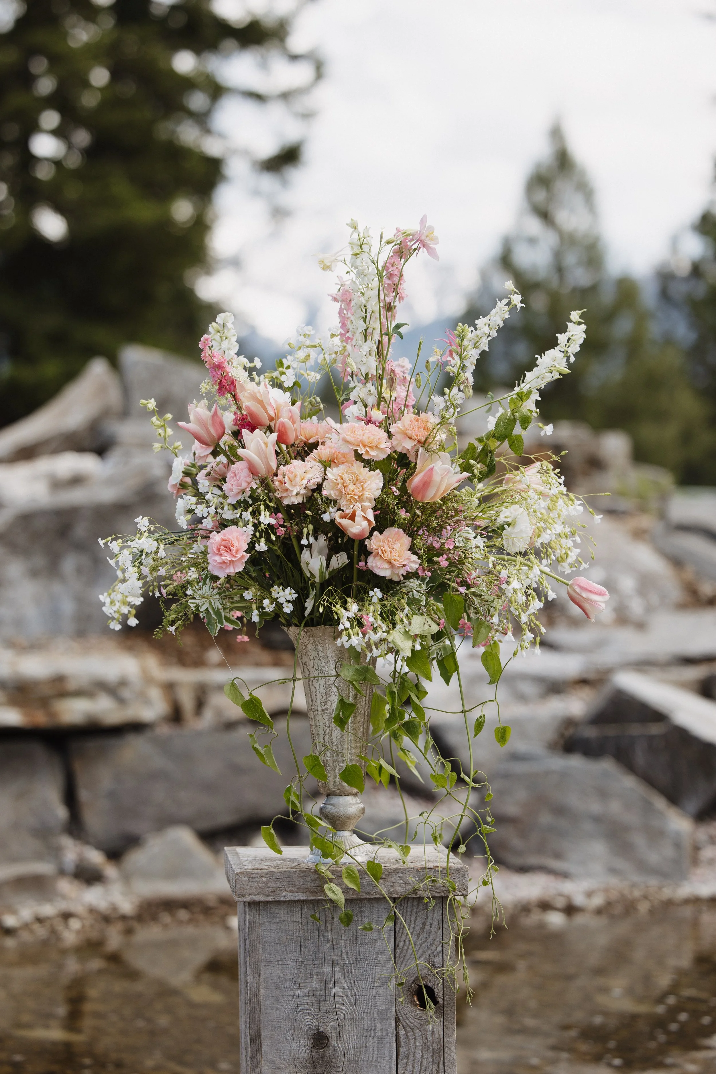 A large arrangement of pink, white, and peach flowers in a decorative vase on a wooden stand outdoors, with rocks and trees in the background.