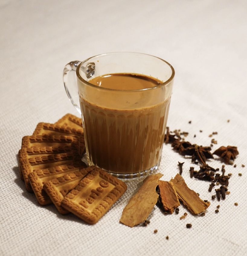 A glass cup filled with chai tea surrounded by cookies, cinnamon sticks, and star anise on a white tablecloth.