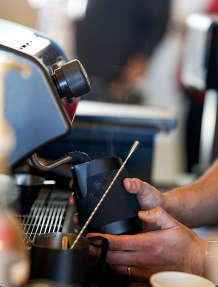 A barista's hands holding a milk frothing pitcher and a coffee steaming machine at a coffee shop.