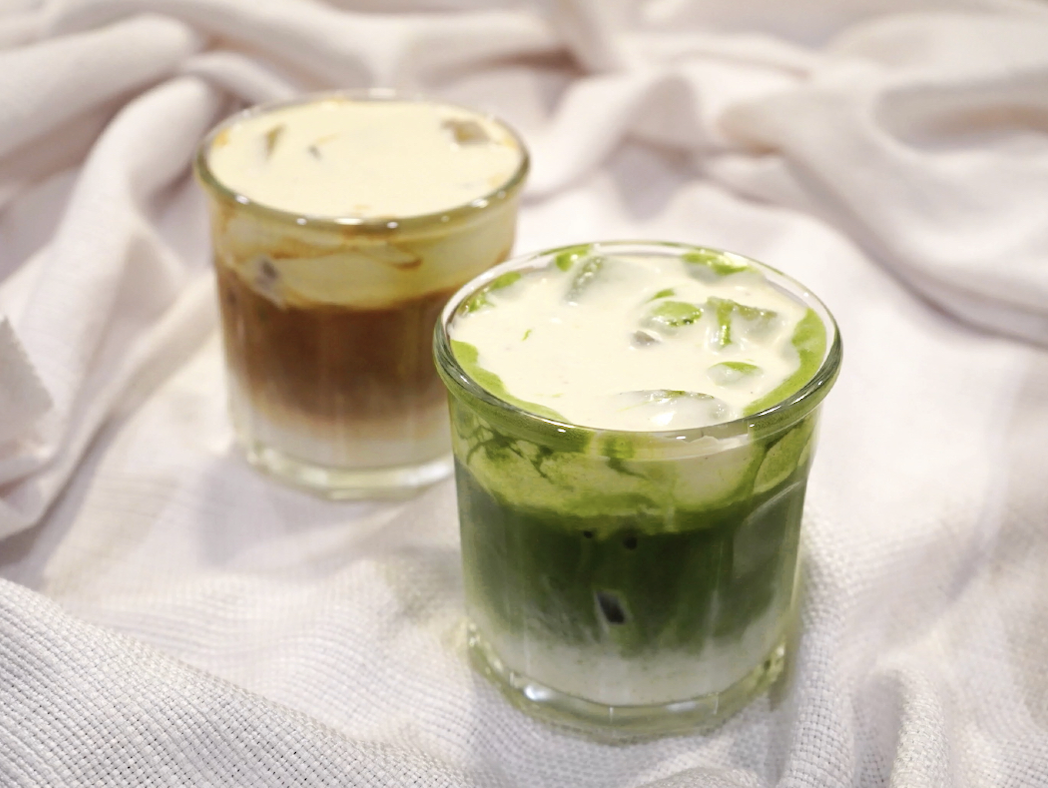 Two glasses of iced coffee, one with a green matcha layer and the other with a brown coffee layer, on a white fabric surface.