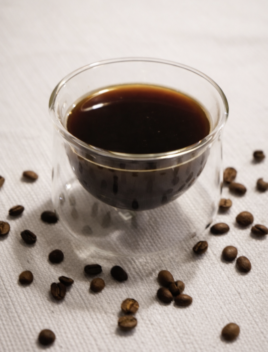 A clear glass cup filled with black coffee on a white textured surface, surrounded by scattered coffee beans.