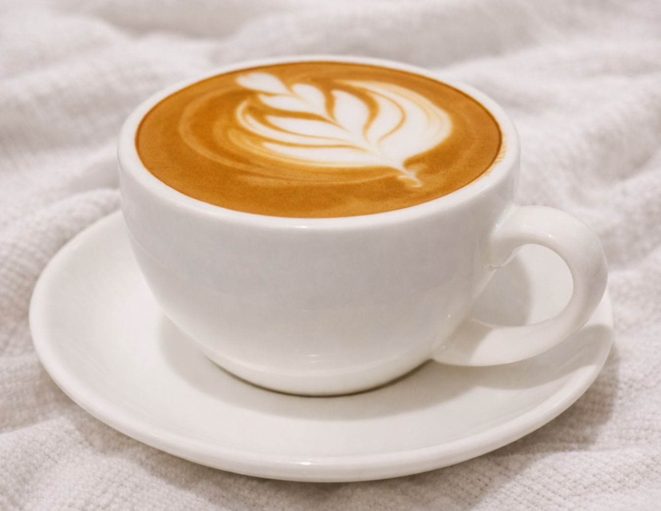 A white coffee cup with a latte art leaf pattern on top, placed on a matching white saucer, on a textured white fabric surface.