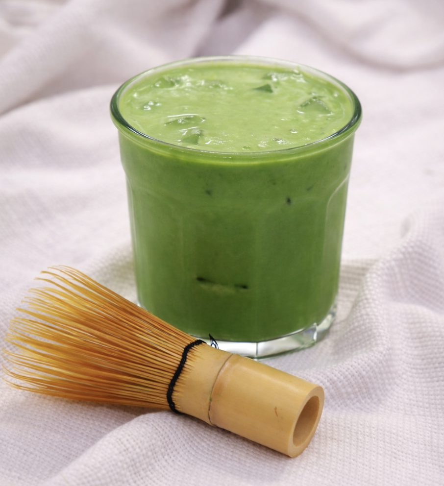 A glass of green matcha latte with a bamboo chasen whisk nearby, on a white cloth background.