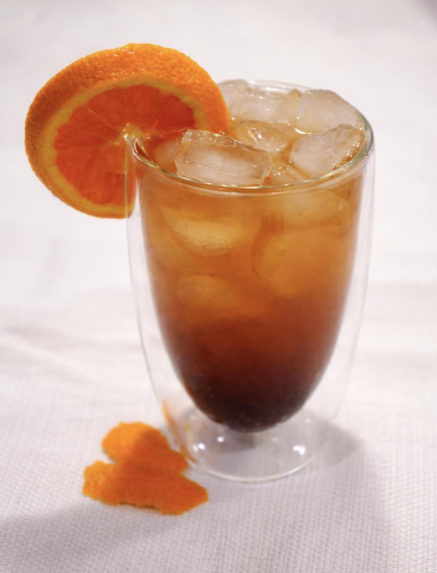A tall glass filled with iced tea, garnished with an orange slice and with some orange peel beside it on a white surface.
