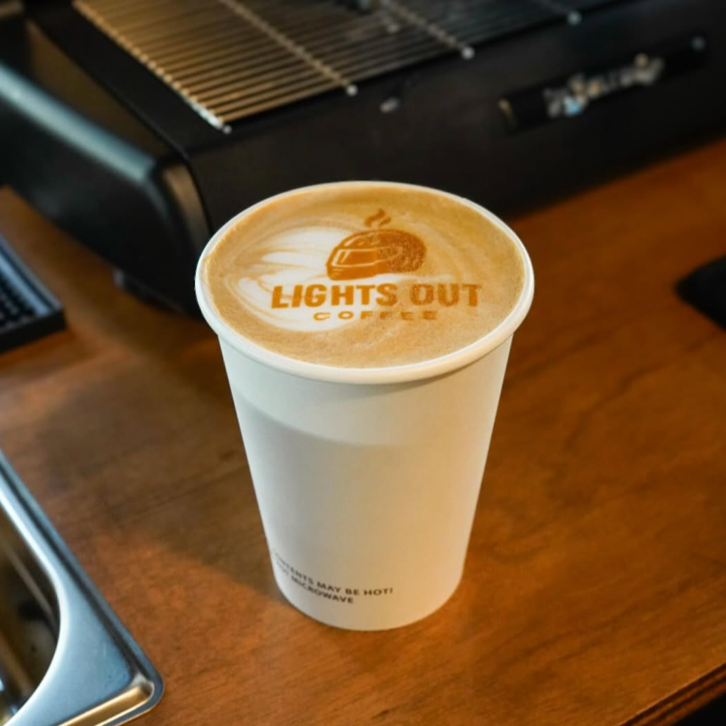 A white coffee cup on a wooden table with latte art that has the logo and text 'Lights Out Coffee' on the foam.