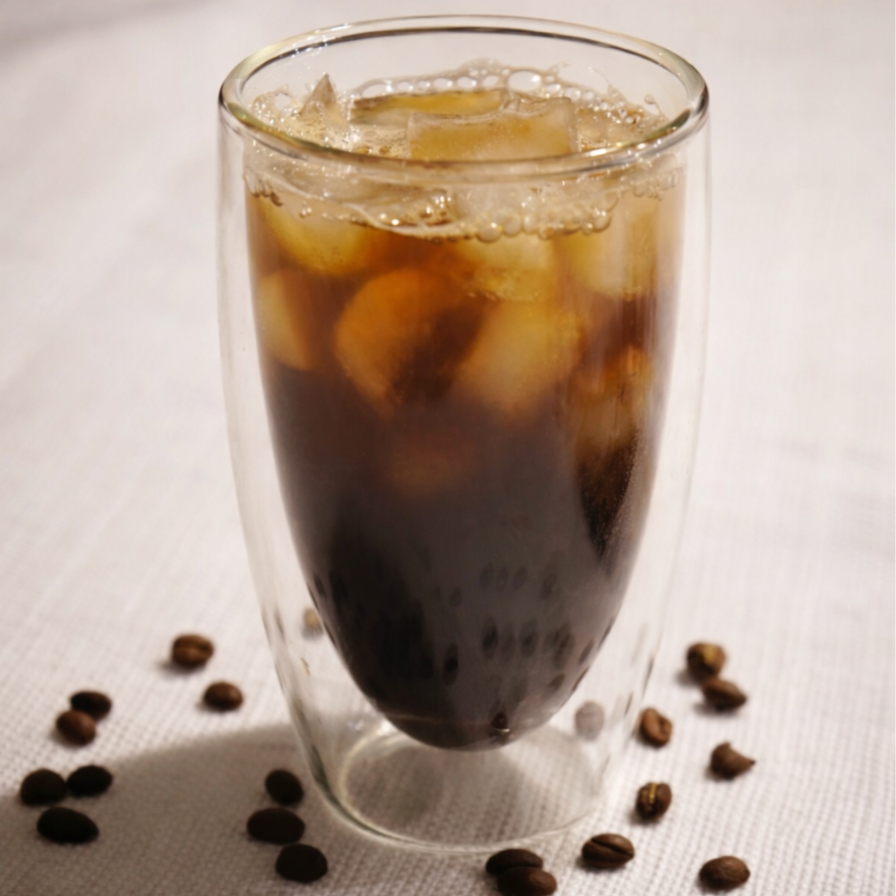 A glass of iced coffee with ice cubes, placed on a white surface with scattered coffee beans around it.
