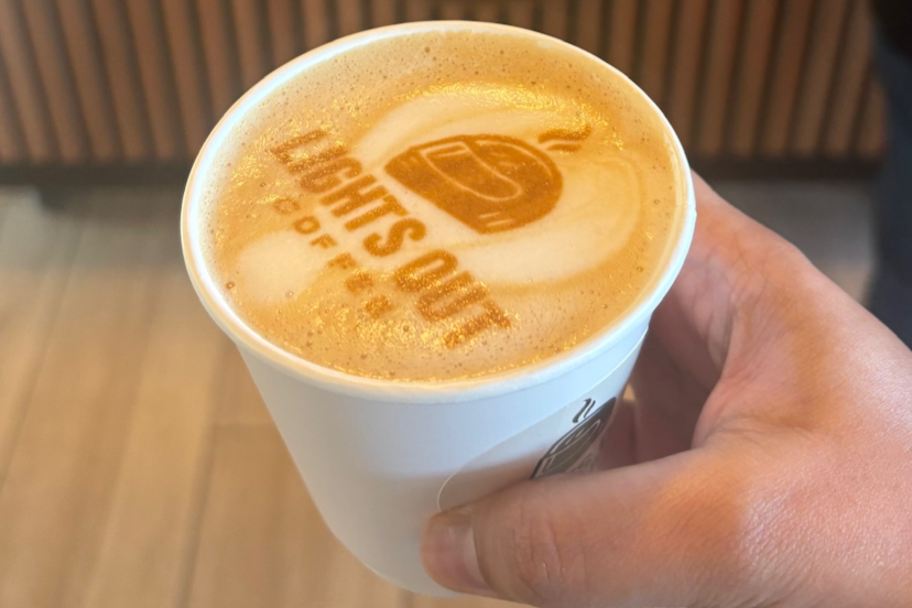 A hand holding a coffee cup with the Starbucks logo on top, featuring latte art and a printed logo on the foam.