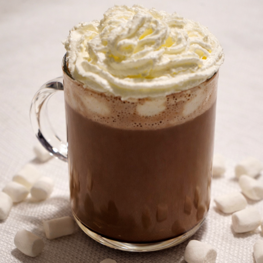 A glass mug of hot chocolate topped with whipped cream, surrounded by marshmallows.