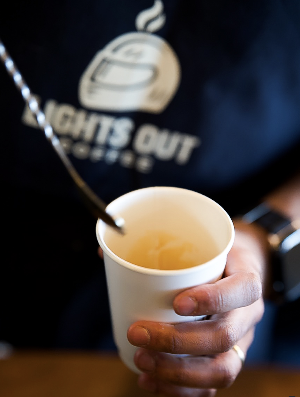 A person holding a white cup filled with tea or coffee, with a silver spoon inside, close-up shot. The person is wearing a black shirt with the logo and text 'CHITS OUT' visible in the background.
