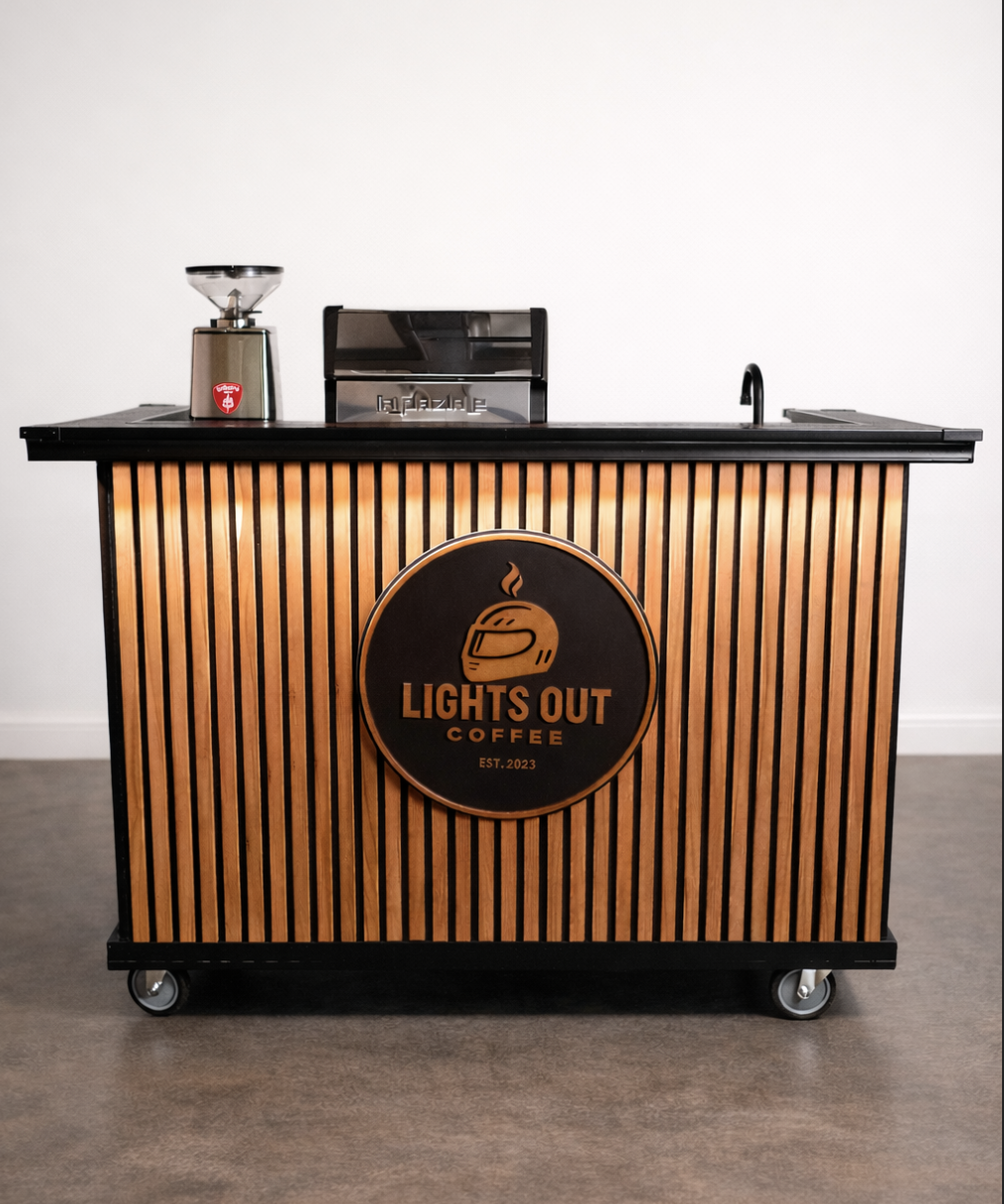 A coffee cart with a wooden slat front and wheels, branded with a "Lights Out Coffee" logo featuring a coffee cup with steam. The cart has a coffee grinder, a juice machine, and a sink on top, set against a plain wall.