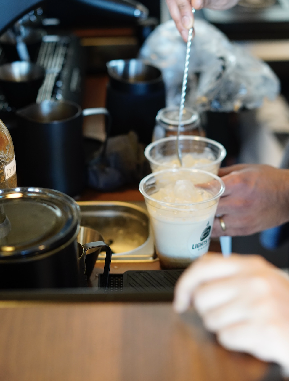 Person stirring a drink with a striped straw at a coffee shop counter, with cups of beverages, a glass jar, and coffee-making equipment visible.