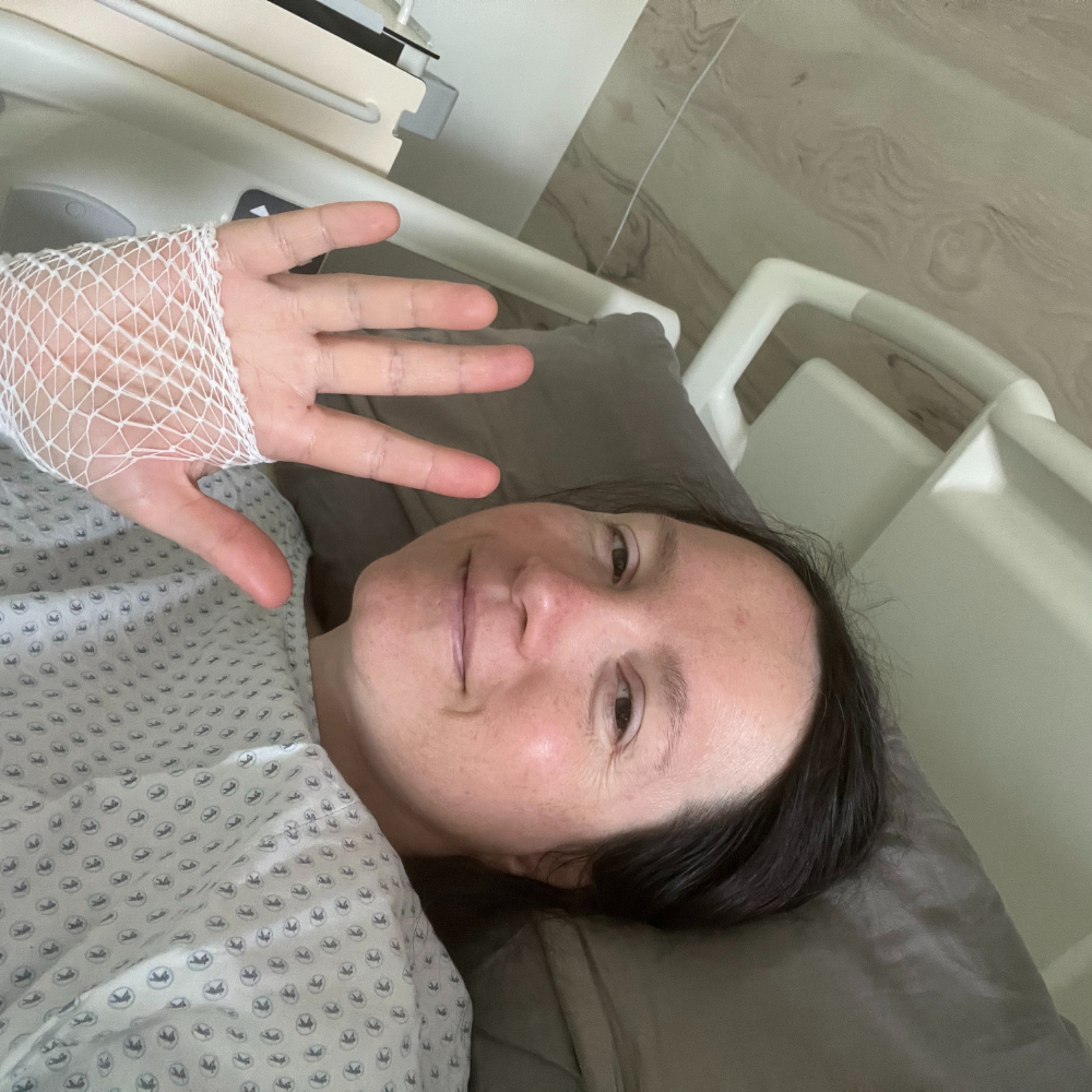 Eine Person liegt im Krankenhausbett, trägt einen medizinischen Gipsverband an der Hand und macht ein Friedenszeichen mit der Hand. Sie lächelt leicht.