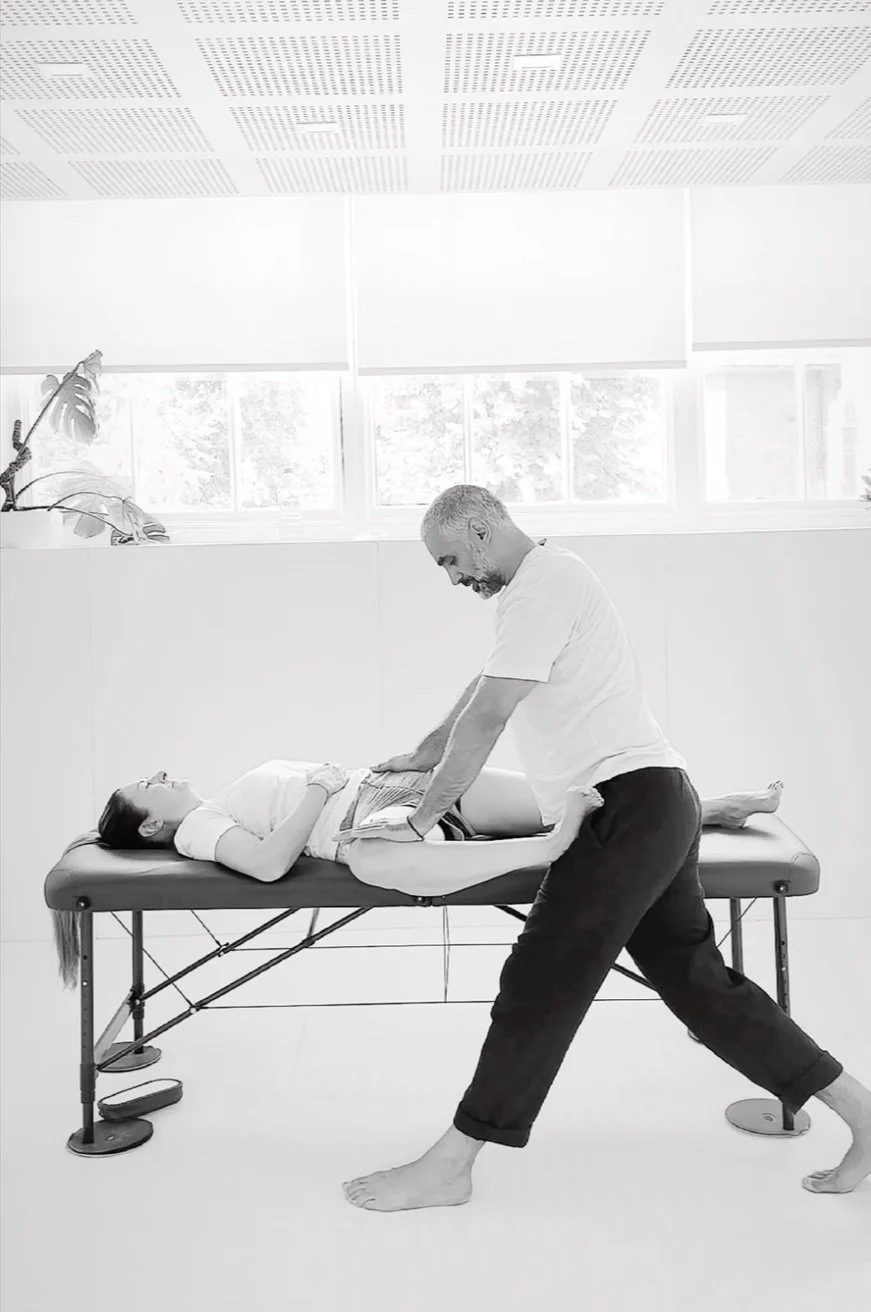 A man providing physiotherapy to a woman lying on a treatment table in a clinic with ample natural light and large windows.