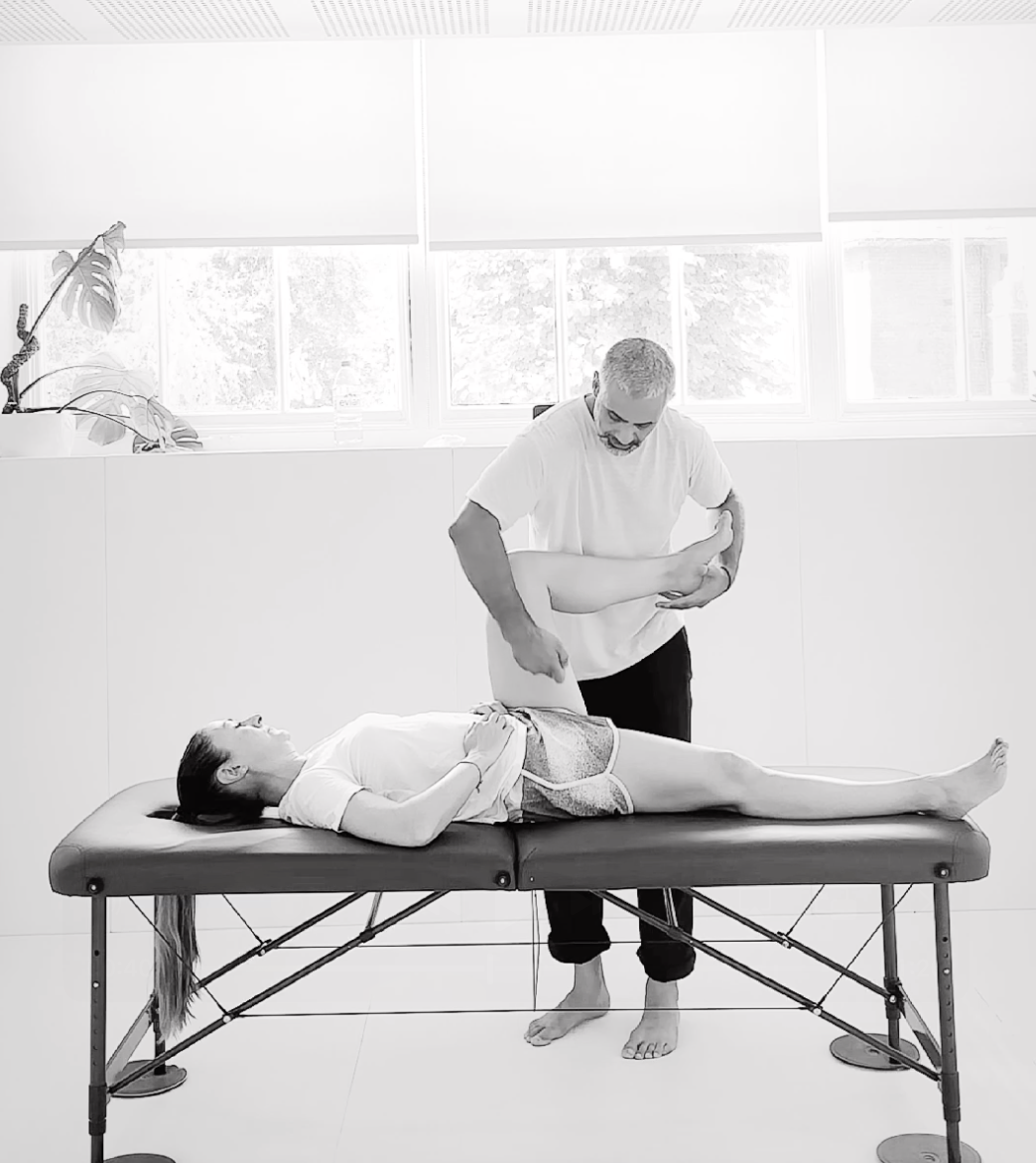 A man gives a massage to a woman lying on a massage table in a brightly lit room with windows and blinds, with a plant on the windowsill.