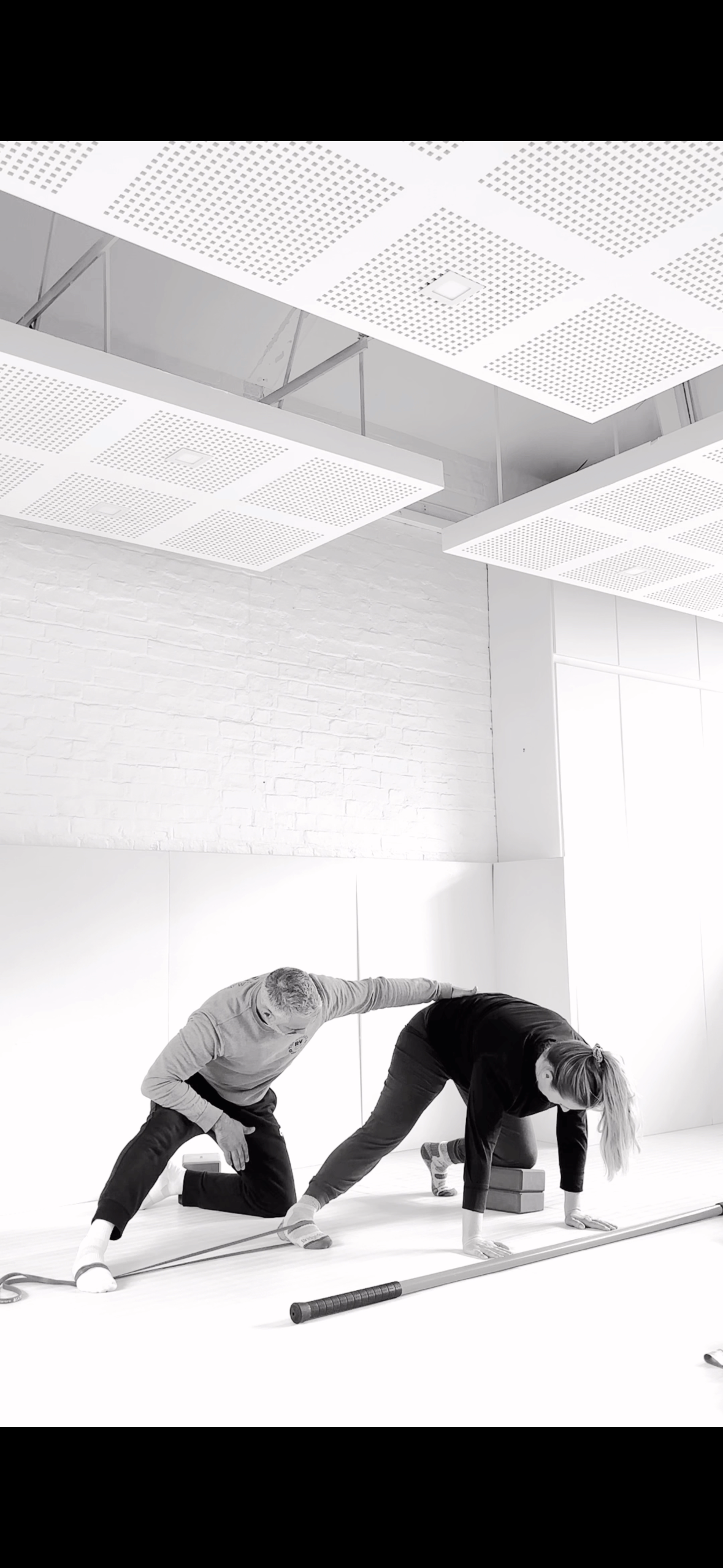 A man and a woman exercising together in a studio, with the woman on her hands and knees and the man assisting her with a stretch.