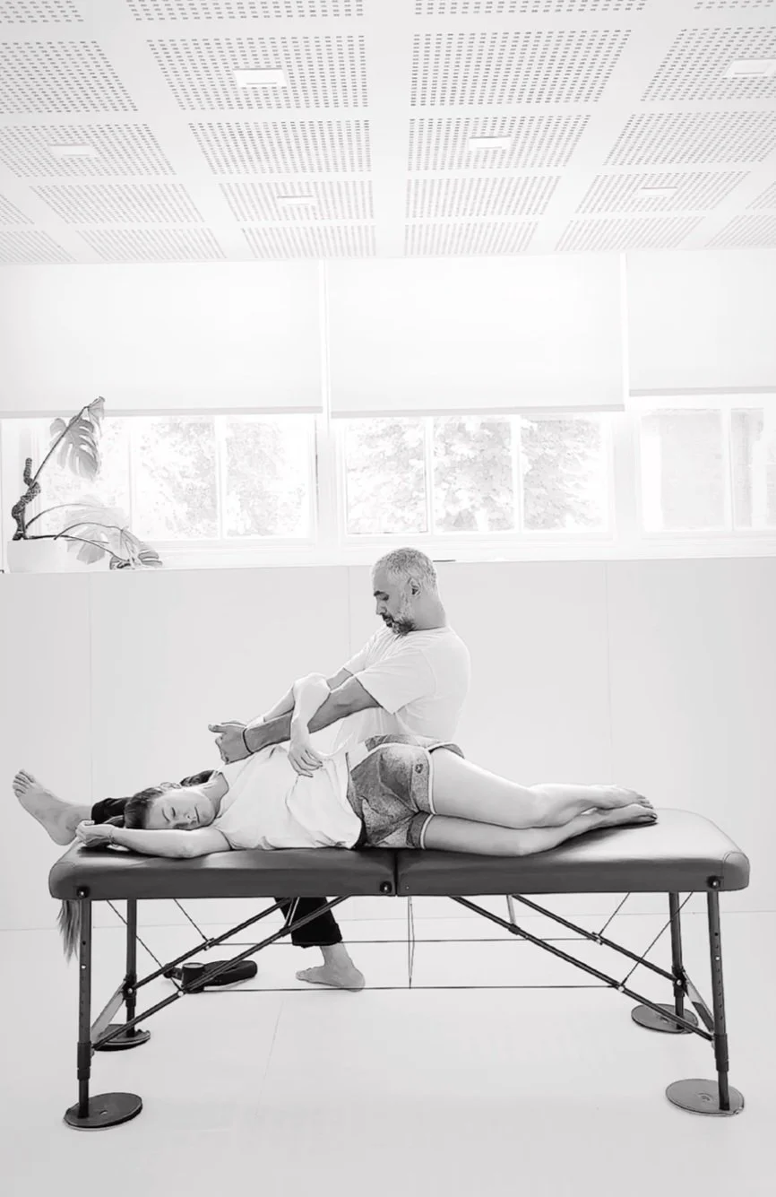 A man providing physical therapy or massage to a woman lying on a treatment table in a bright room with large windows and ceiling lights.