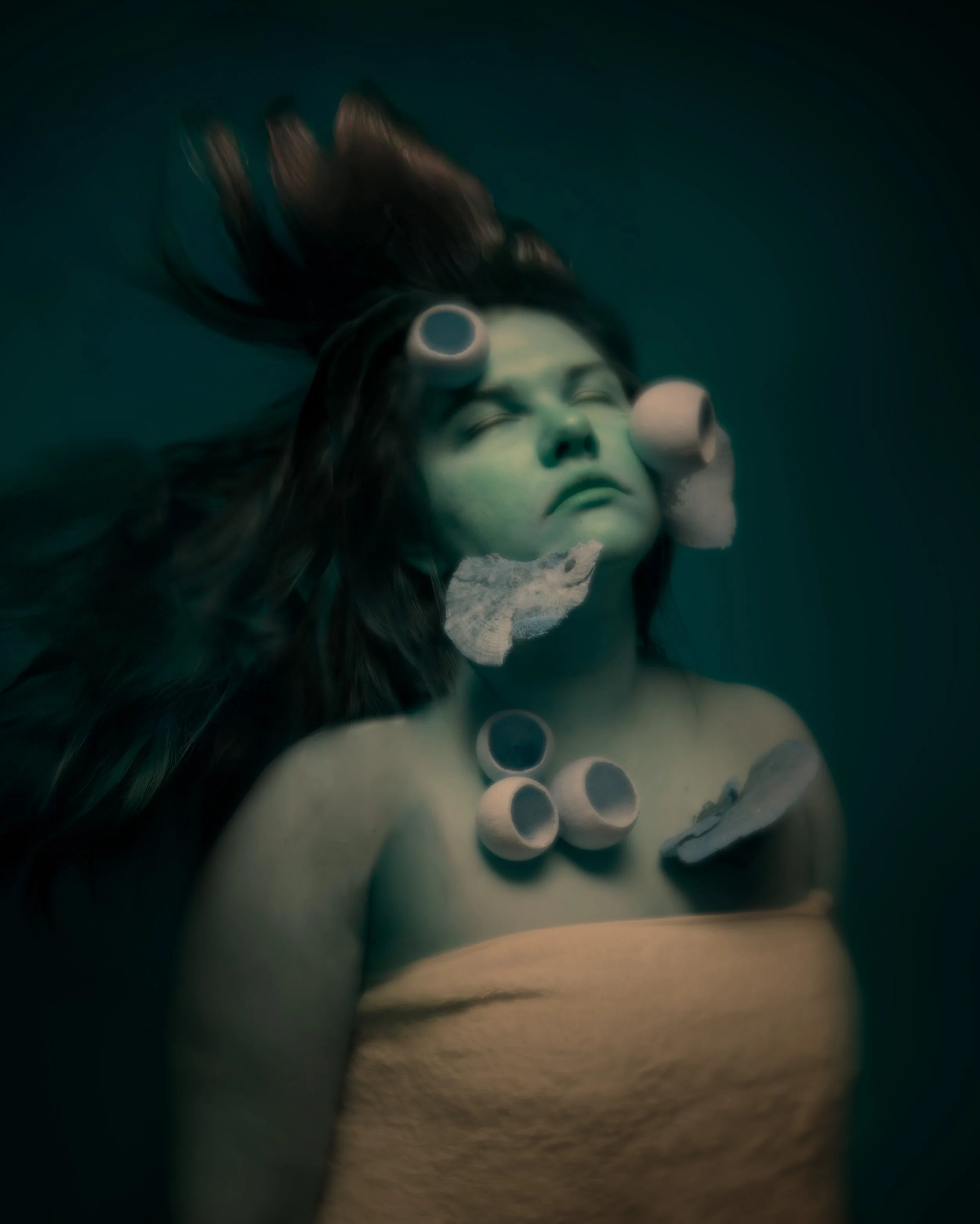A woman with dark hair is underwater, her eyes closed, with bubbles and rocks around her face and shoulders.