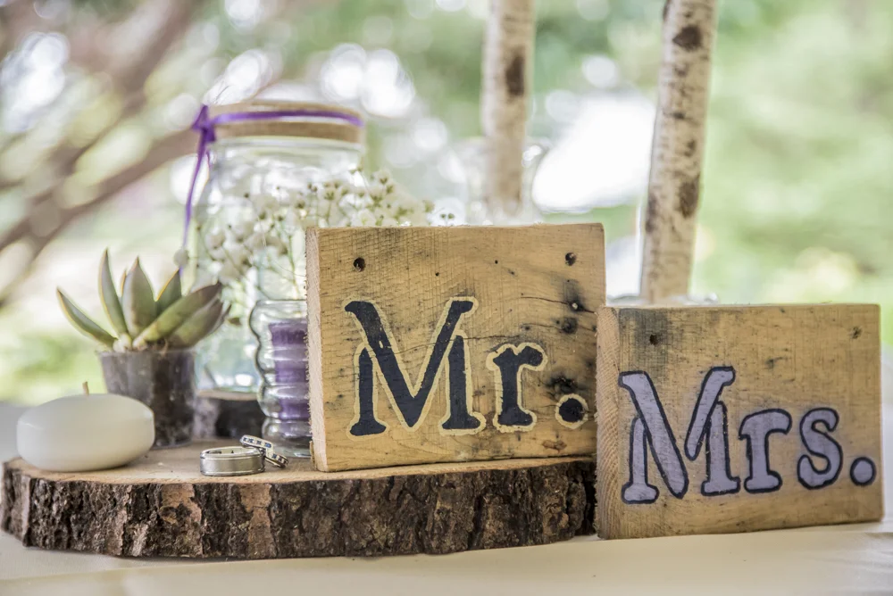 Wooden signs with 'Mr.' and 'Mrs.' written on them, placed on a tree stump with small plants, a glass jar filled with white flowers, a white candle, and rings, outdoors with blurred trees in the background.