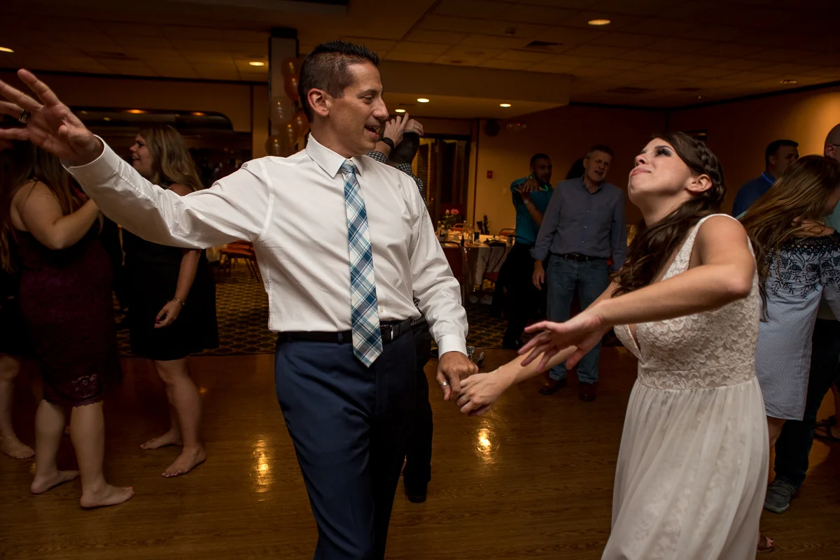 A man and woman dancing happily at a party, holding hands, with other guests dancing in the background.