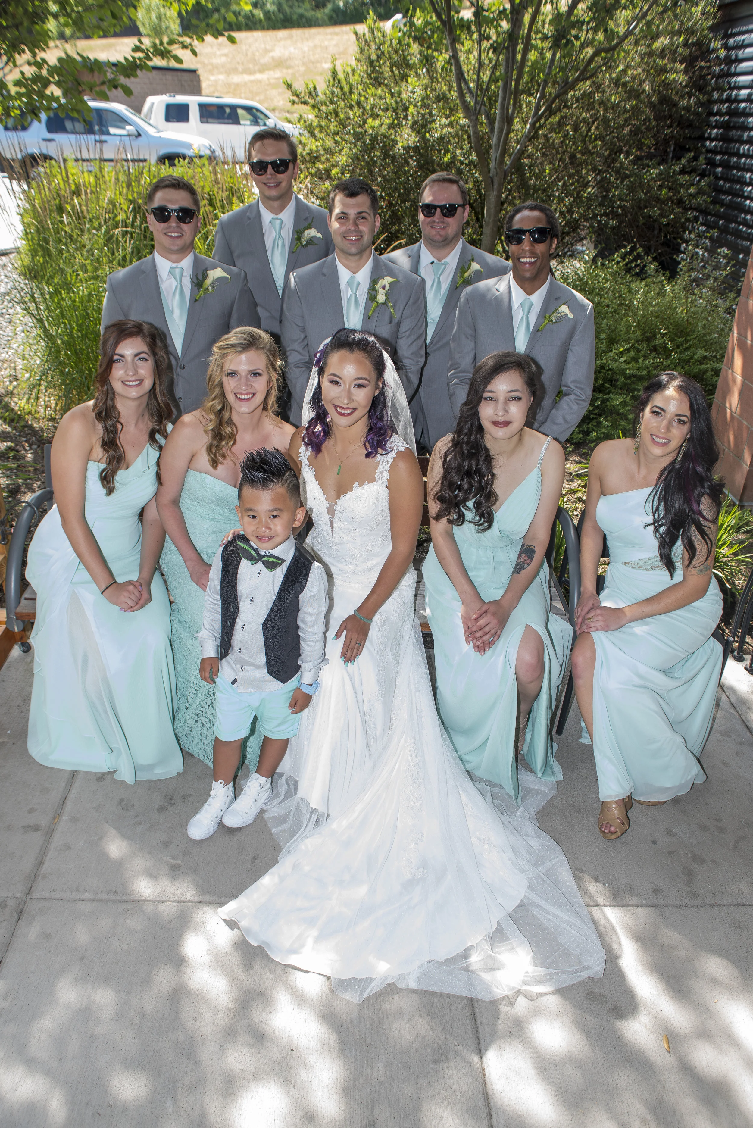 Group photo of wedding party outdoors. The bride in a white gown, surrounded by bridesmaids in light blue dresses, and groomsmen in gray suits with light blue ties and boutonnieres. A young boy in front wears a vest, bow tie, and shorts. Background i