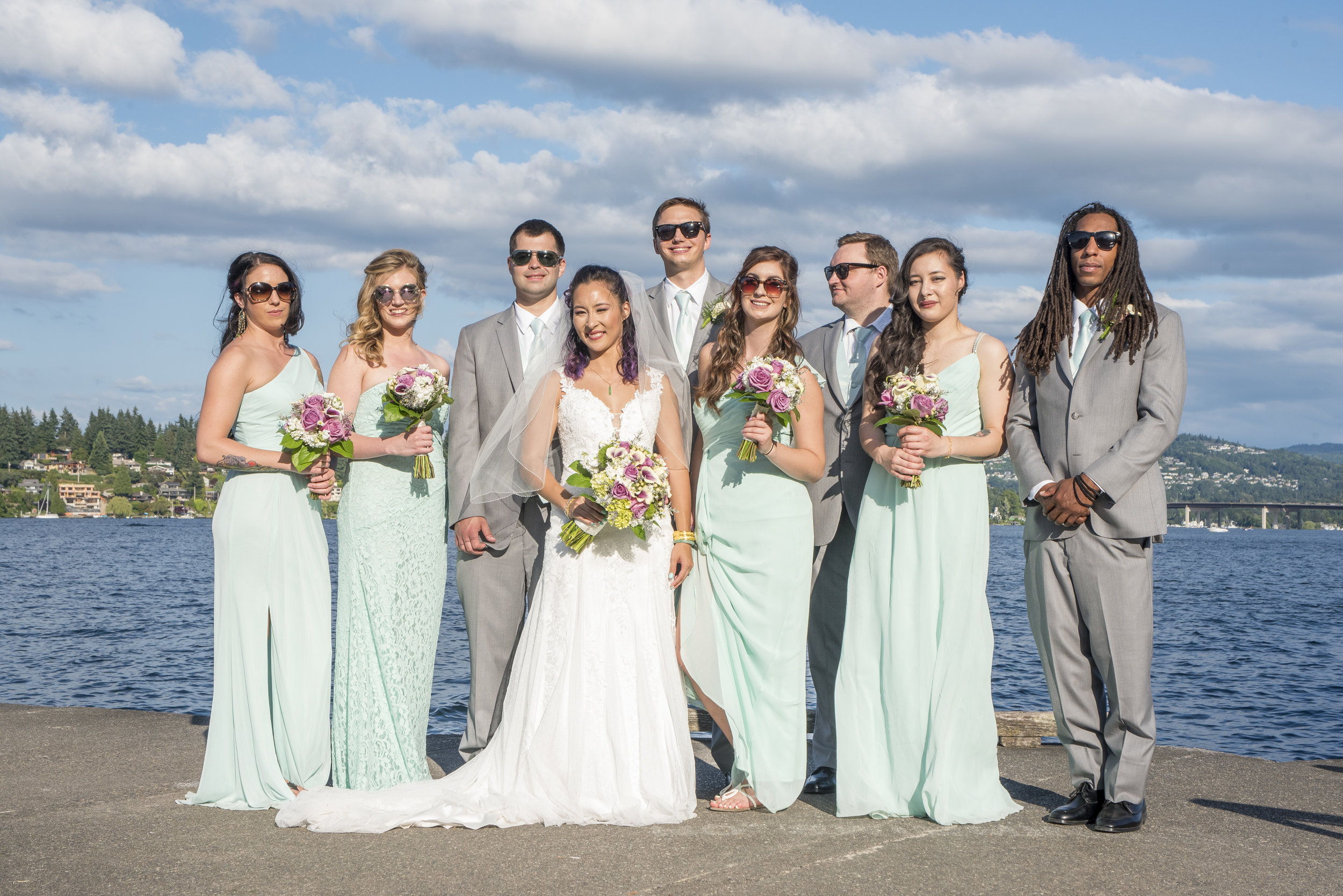 A group of ten people, including a bride and groom, standing by a body of water with a bridge and hillside in the background on a sunny day.