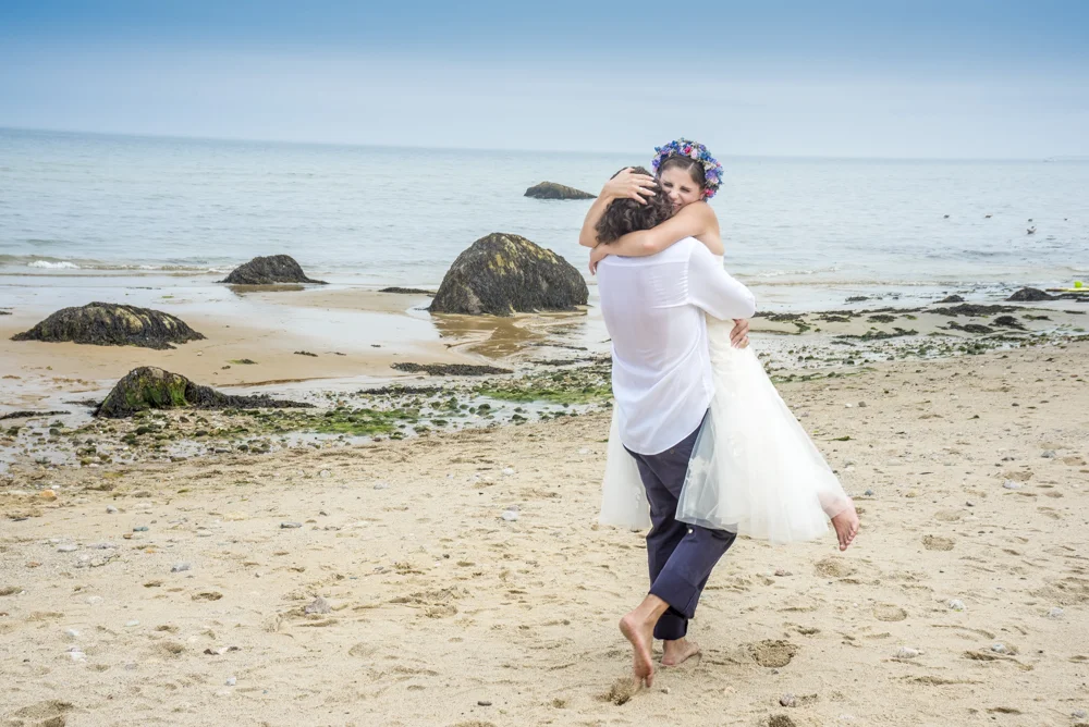 A couple hugging on the beach, with the woman in a white dress and flower crown, and the man in a white shirt and dark pants, both barefoot on the sand. Rocks and the ocean are in the background.