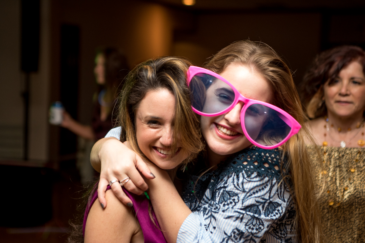 Two women hugging and smiling at a social gathering, one wearing large pink sunglasses.