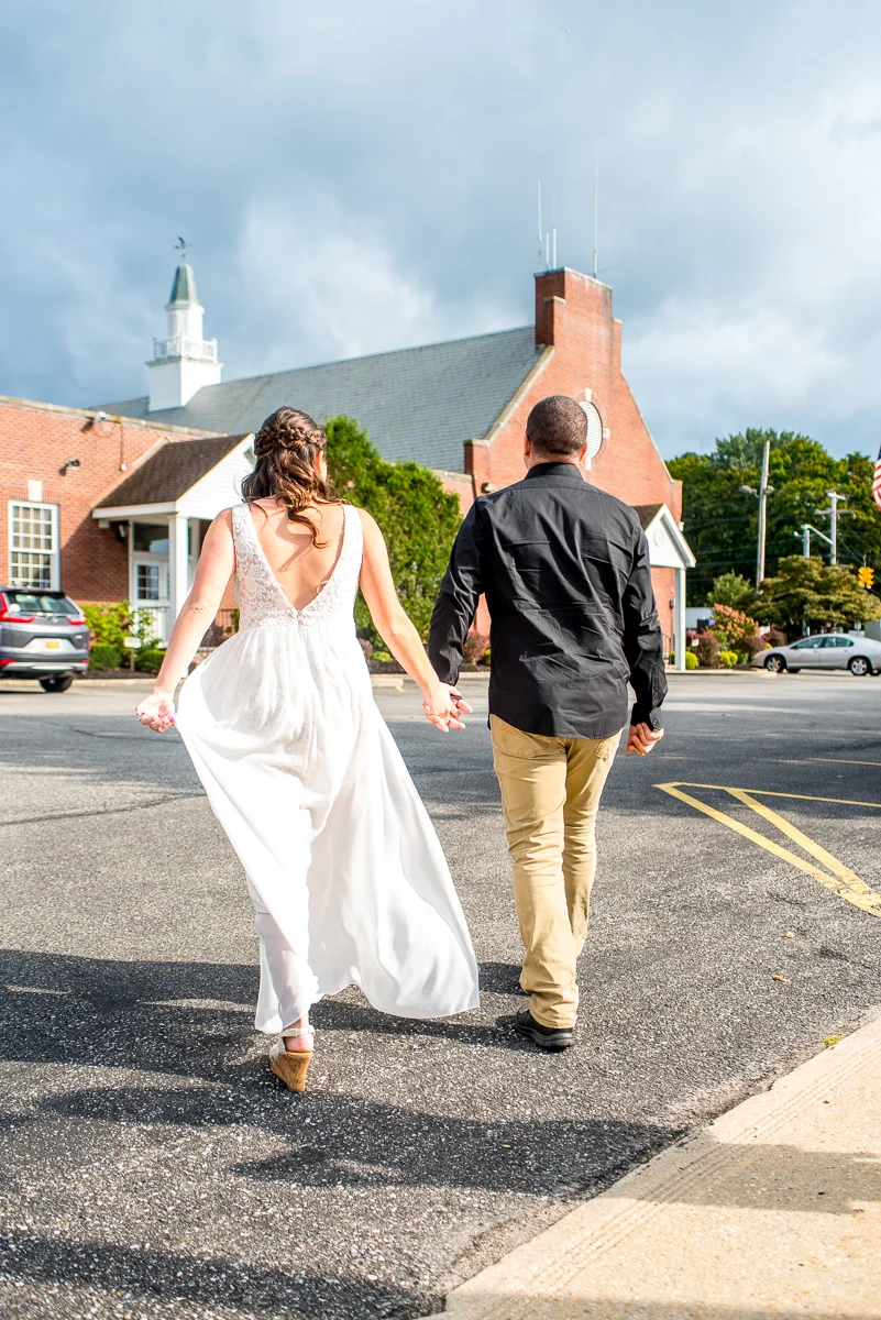 A couple holding hands and walking away from the camera in a parking lot, with a church and trees in the background. The woman is wearing a white dress and the man is wearing a black shirt and tan pants.