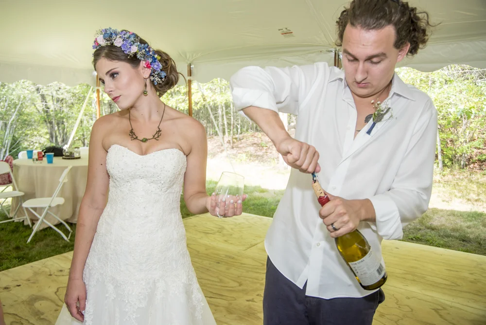 A woman in a white wedding dress and floral crown stands next to a man opening a bottle of wine at a reception tent outdoors.