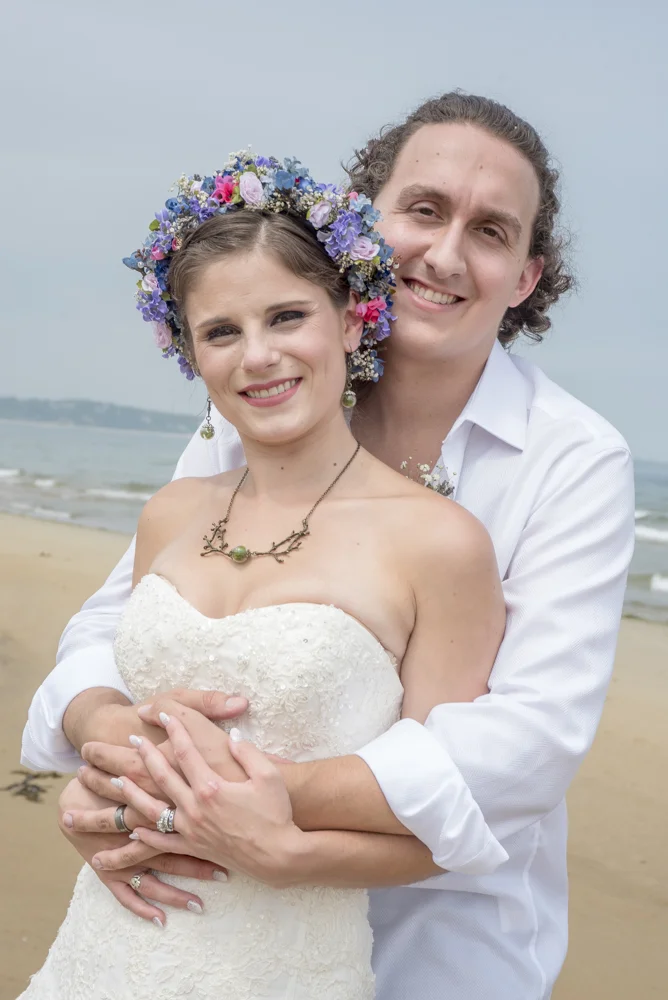 A happy couple on a beach, with the woman wearing a white wedding dress and floral headpiece, and the man in a white shirt, embracing each other and smiling at the camera.