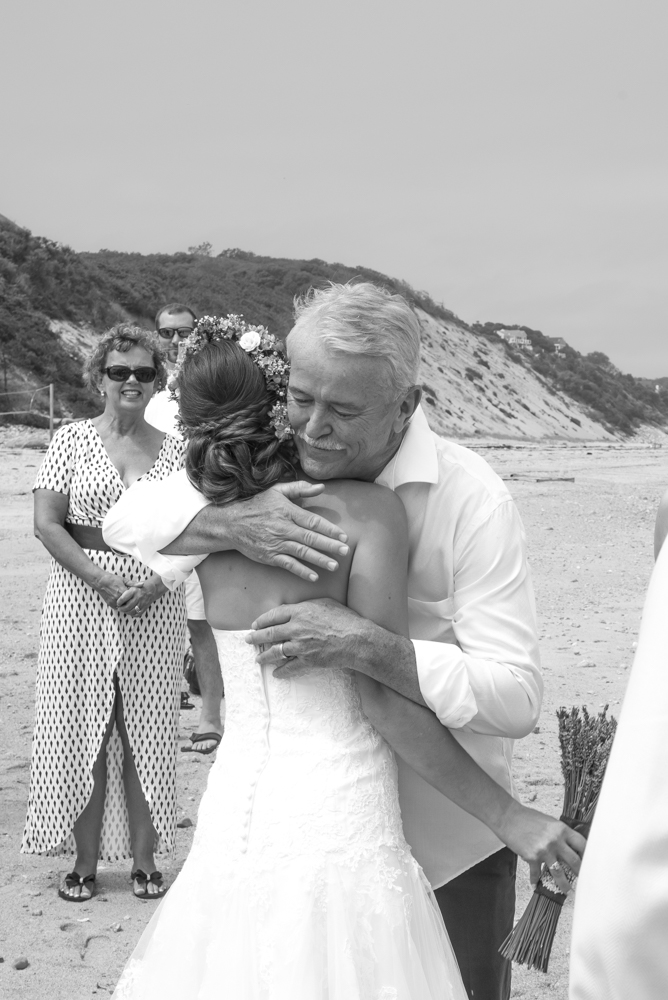 A father and daughter share a hug on the beach during her wedding, with guests in the background.