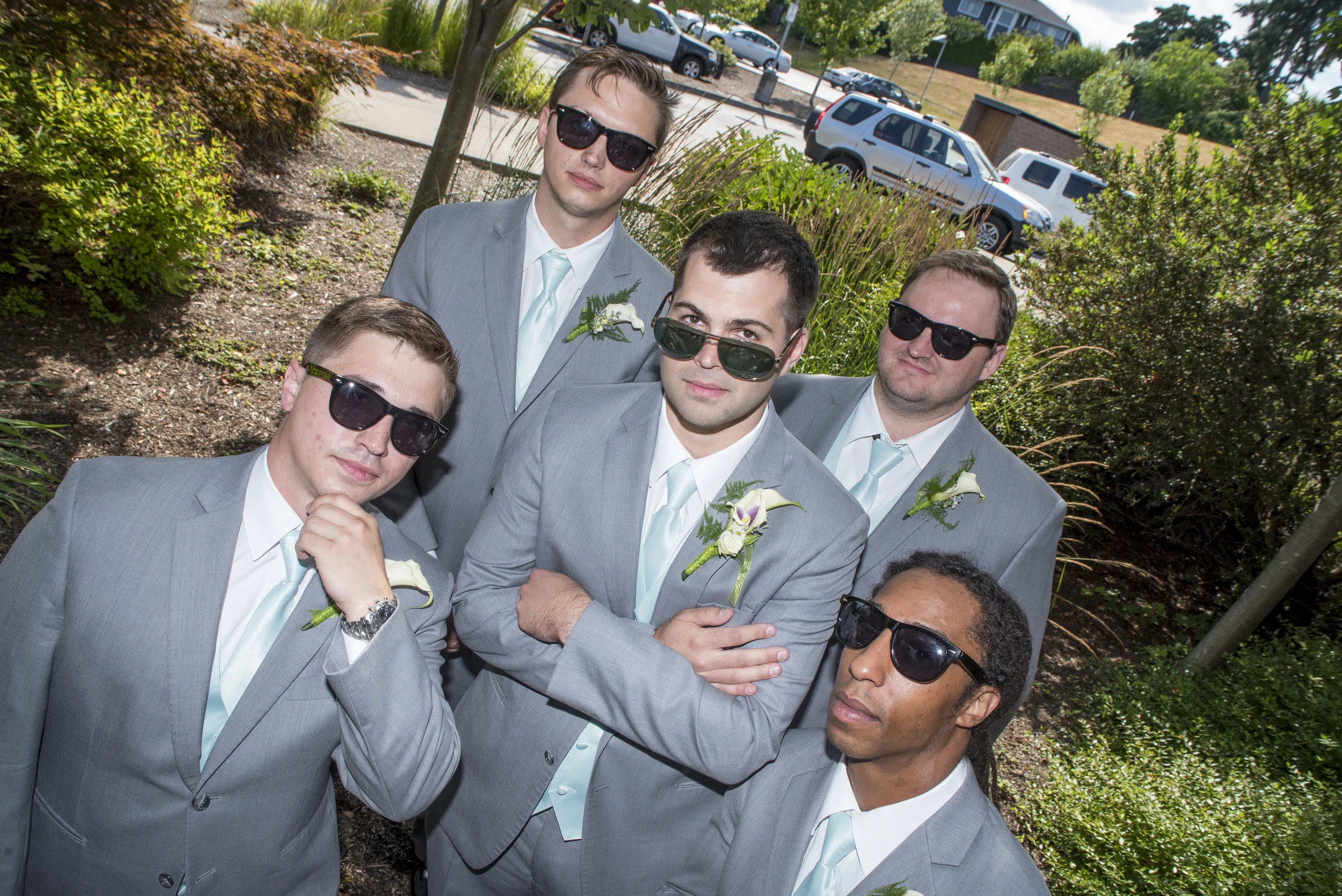 A group of six men in gray suits with light blue ties, wearing sunglasses, standing outdoors among greenery and shrubs, posing for a photo. Some men have white flowers pinned to their suits.